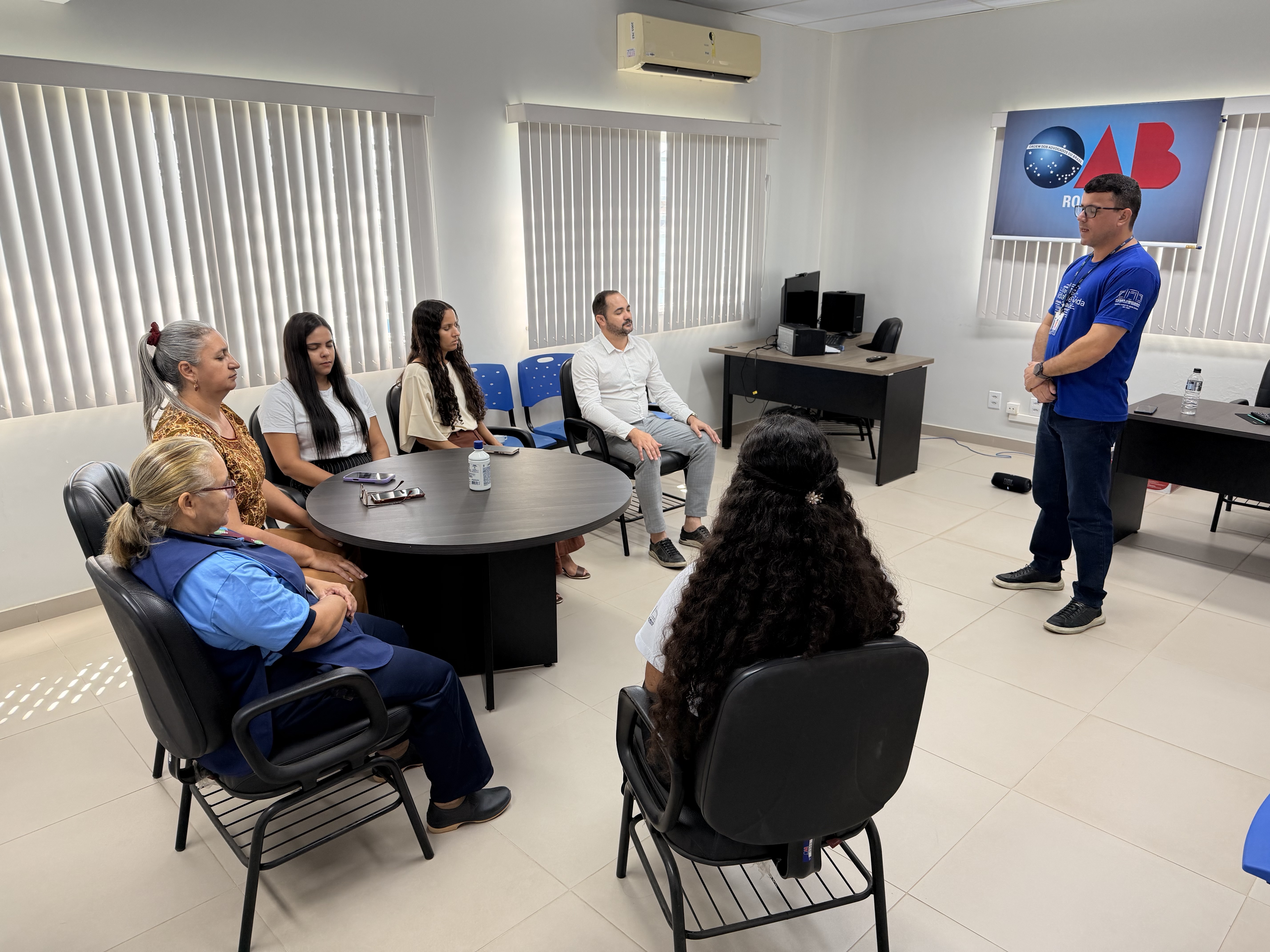 Um instrutor em pé conversa com um grupo de pessoas sentadas em círculo em uma sala de reuniões.