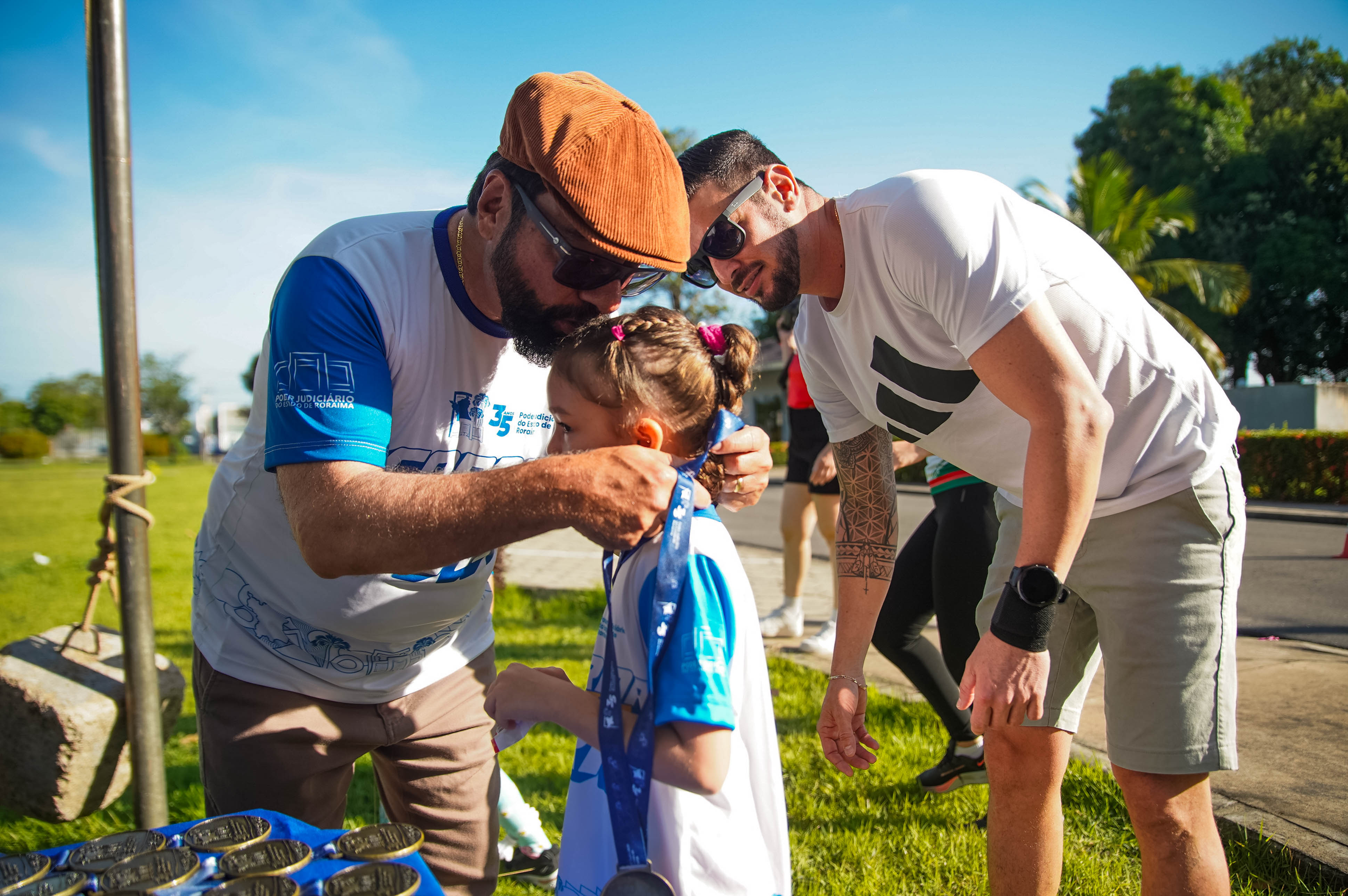 Imagem colorida registra uma criança recebendo a medalha da Corrida Kids. O presidente do TJRR, desembargador Leonardo Cupello, coloca a medalha no pescoço da menina, enquanto outro homem acompanha o momento, em área externa do Complexo Sociocultural. Imagem colorida registra uma criança recebendo a medalha da Corrida Kids. O presidente do TJRR, desembargador Leonardo Cupello, coloca a medalha no pescoço da menina, enquanto outro homem acompanha o momento, em área externa do Complexo Sociocultural.