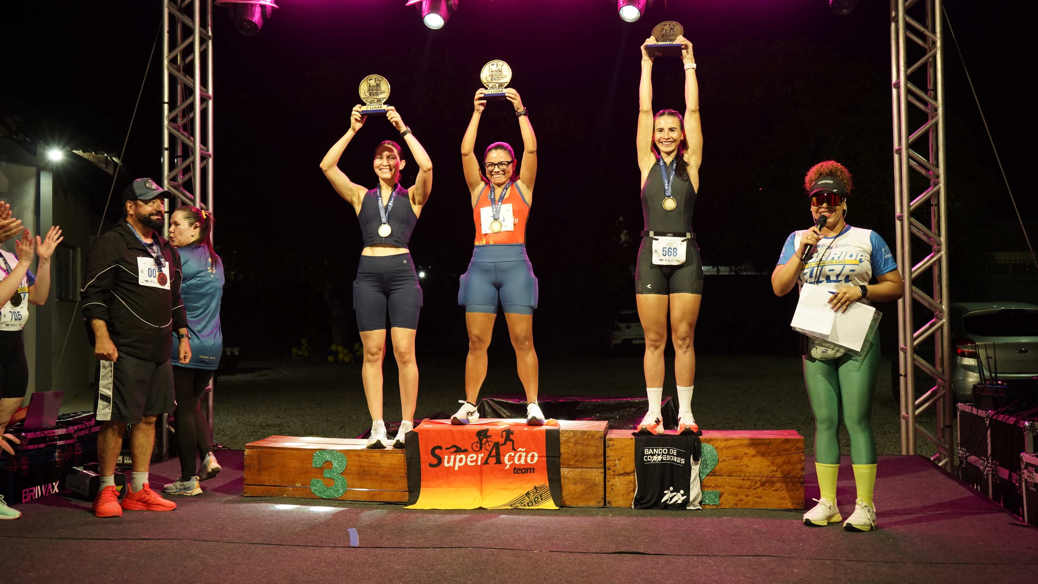 Imagem colorida mostra três atletas no pódio feminino da Corrida TJRR 35 Anos, segurando troféus acima da cabeça e usando medalhas. Ao lado, integrantes da organização acompanham a premiação no palco iluminado. Imagem colorida mostra três atletas no pódio feminino da Corrida TJRR 35 Anos, segurando troféus acima da cabeça e usando medalhas. Ao lado, integrantes da organização acompanham a premiação no palco iluminado.
