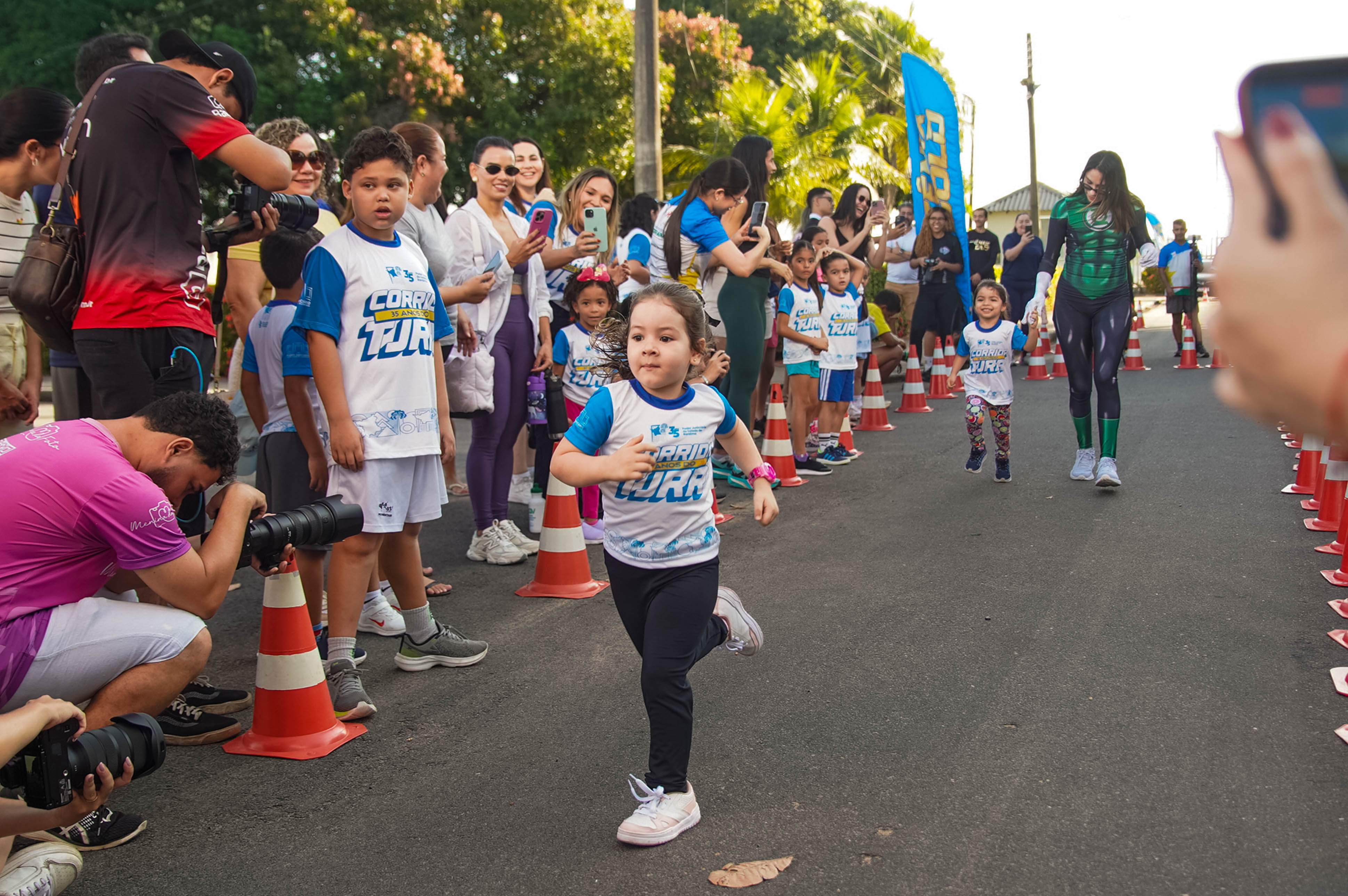 Imagem colorida mostra crianças participando da Corrida Kids em percurso delimitado por cones. Uma menina corre em primeiro plano, enquanto familiares, fotógrafos e outros participantes acompanham e registram a prova nas laterais. Imagem colorida mostra crianças participando da Corrida Kids em percurso delimitado por cones. Uma menina corre em primeiro plano, enquanto familiares, fotógrafos e outros participantes acompanham e registram a prova nas laterais.