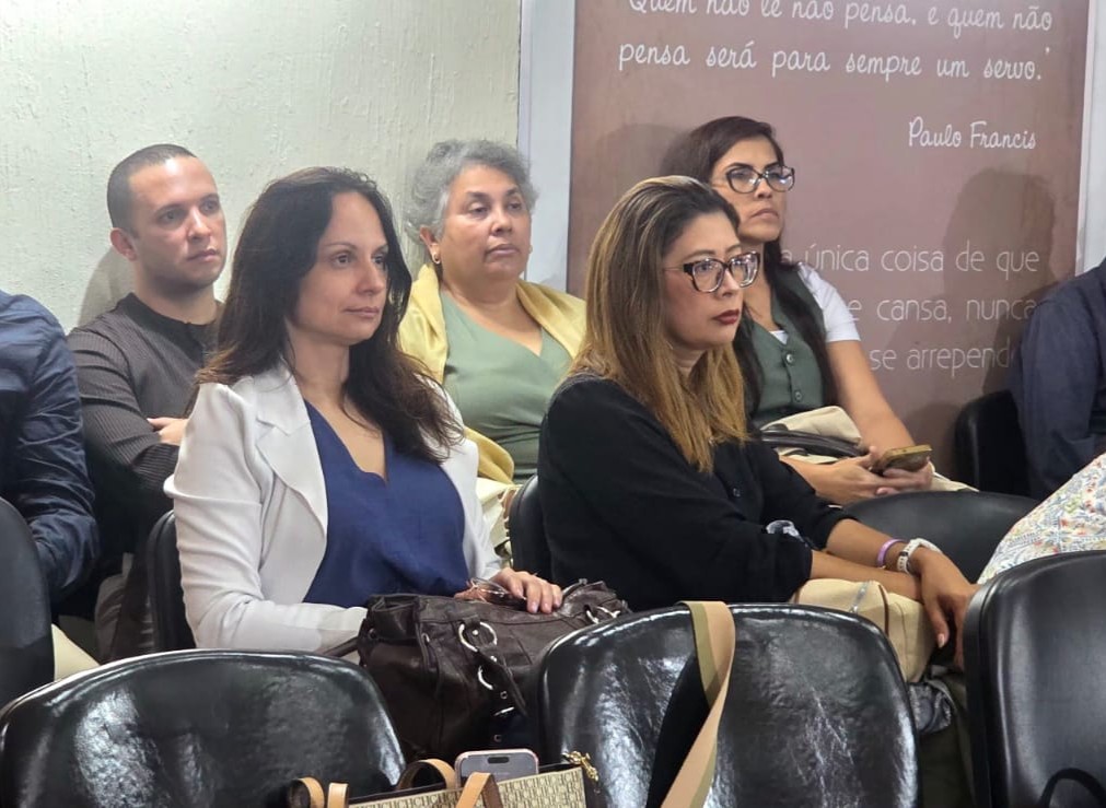 Fotografia de pessoas sentadas em cadeiras dentro de uma sala. Em primeiro plano, duas mulheres aparecem sentadas, enquanto outras pessoas estão ao fundo. Na parede, há uma frase de Paulo Freire escrita em letras brancas.