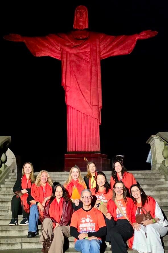 Fotografia noturna de um grupo de pessoas reunidas nas escadarias do Cristo Redentor, no Rio de Janeiro. Ao fundo, a estátua do Cristo está iluminada na cor vermelha.