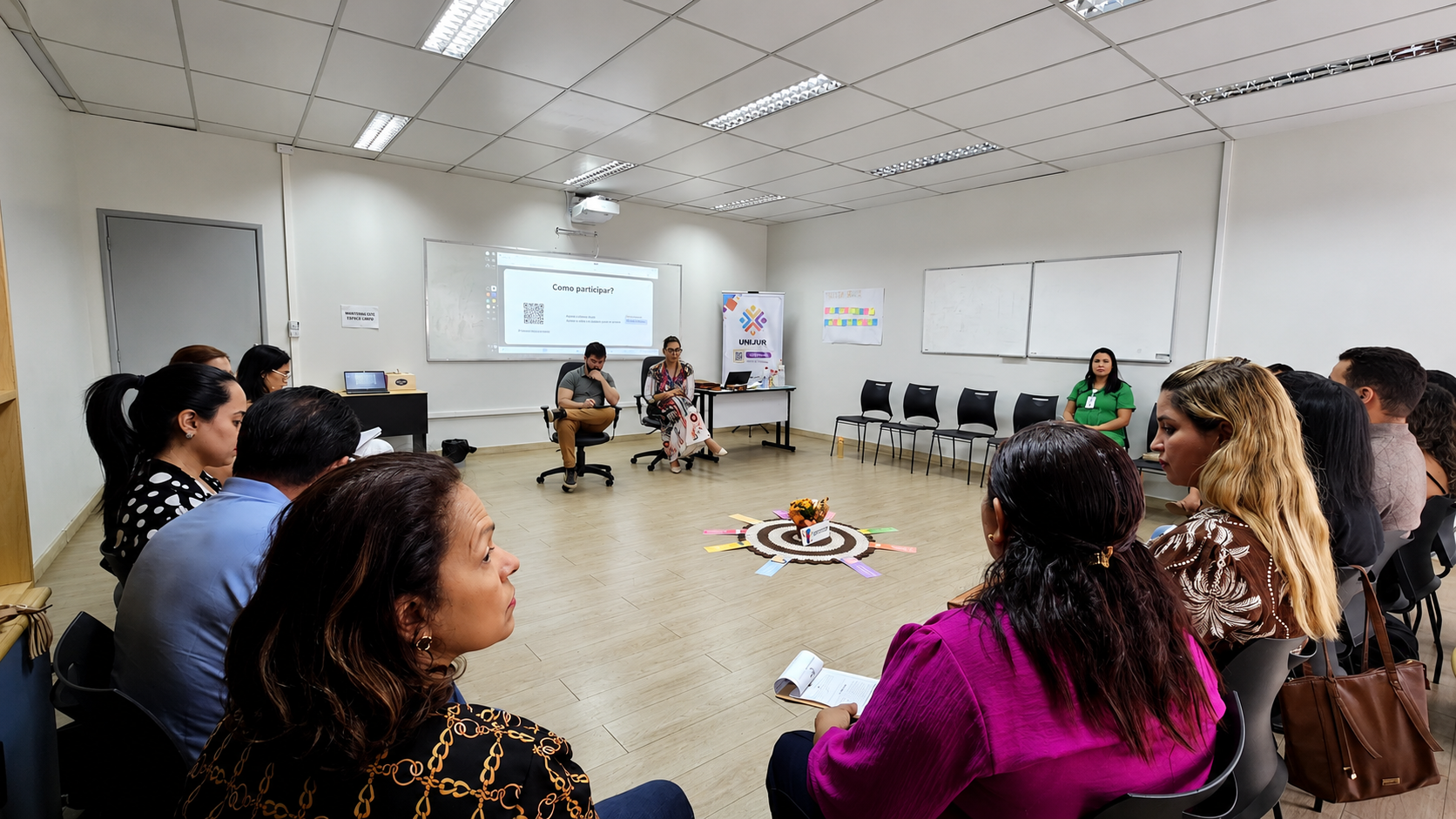 Imagem colorida registra visão ampla da formação em Justiça Restaurativa, com os participantes sentados em círculo e facilitadores conduzindo a atividade à frente da sala. Ao fundo, uma apresentação é projetada na parede com orientações para participação na dinâmica. Imagem colorida registra visão ampla da formação em Justiça Restaurativa, com os participantes sentados em círculo e facilitadores conduzindo a atividade à frente da sala. Ao fundo, uma apresentação é projetada na parede com orientações para participação na dinâmica.