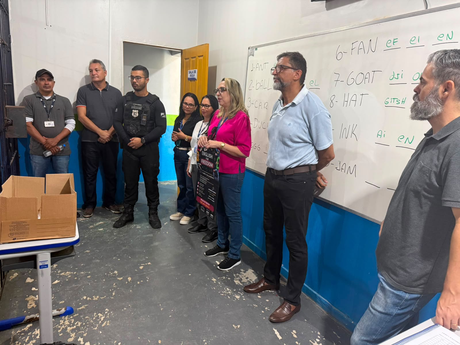 Imagem colorida mostra um grupo de pessoas em pé dentro de uma sala de aula de parede branca e azul. Uma mulher de blusa rosa segura um cartaz que diz "ENGLISH FOR FREEDOM" em frente a um quadro branco com palavras em inglês e pronúncias anotadas. Imagem colorida mostra um grupo de pessoas em pé dentro de uma sala de aula de parede branca e azul. Uma mulher de blusa rosa segura um cartaz que diz "ENGLISH FOR FREEDOM" em frente a um quadro branco com palavras em inglês e pronúncias anotadas.