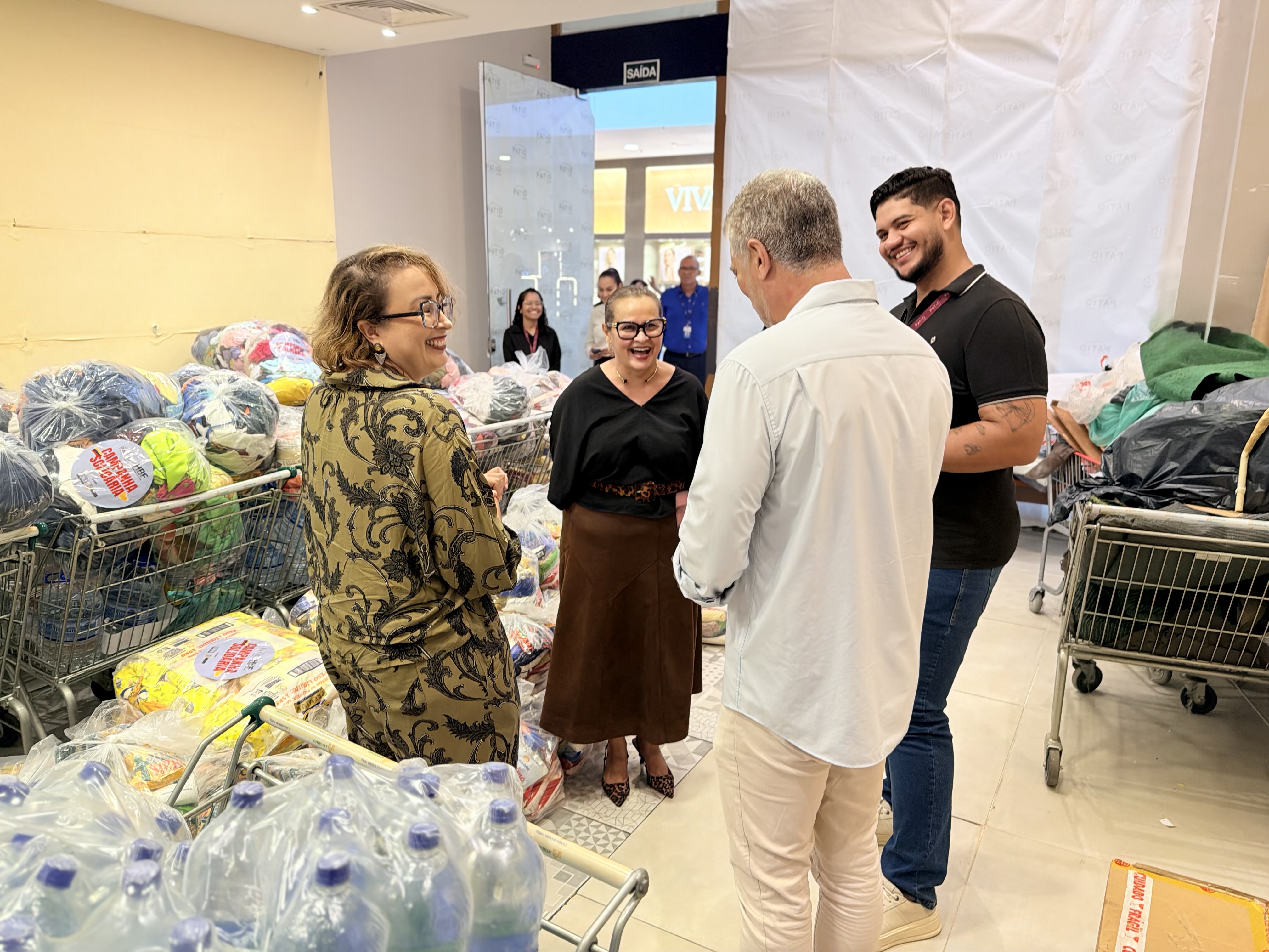 Imagem colorida mostra quatro pessoas conversando em tom descontraído e sorrindo dentro de um ambiente amplo. Em volta do grupo, há diversos carrinhos de supermercado e sacos cheios de doações como alimentos, roupas e garrafas de água. Imagem colorida mostra quatro pessoas conversando em tom descontraído e sorrindo dentro de um ambiente amplo. Em volta do grupo, há diversos carrinhos de supermercado e sacos cheios de doações como alimentos, roupas e garrafas de água.