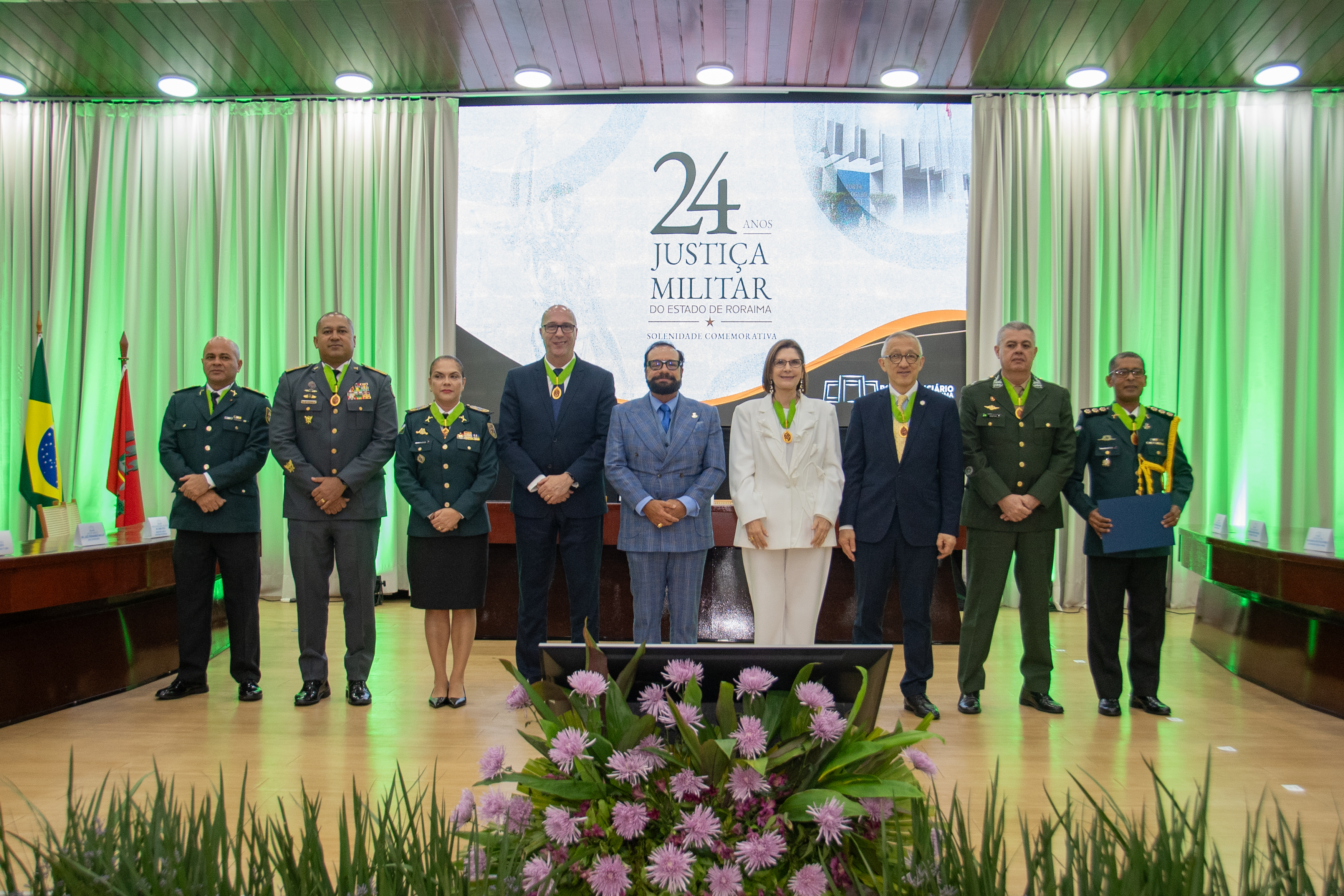 Imagem colorida registra autoridades e homenageados da solenidade comemorativa dos 24 anos da Justiça Militar de Roraima posando para fotografia oficial no palco. Ao centro está o presidente do TJRR, desembargador Leonardo Cupello, acompanhado da presidente do Superior Tribunal Militar, ministra Maria Elizabeth Guimarães, de representantes dos Tribunais de Justiça Militar e de integrantes das corporações militares. Ao fundo, o painel exibe a identidade visual dos 24 anos da Justiça Militar do Estado de Roraima, com iluminação verde no ambiente. Imagem colorida registra autoridades e homenageados da solenidade comemorativa dos 24 anos da Justiça Militar de Roraima posando para fotografia oficial no palco. Ao centro está o presidente do TJRR, desembargador Leonardo Cupello, acompanhado da presidente do Superior Tribunal Militar, ministra Maria Elizabeth Guimarães, de representantes dos Tribunais de Justiça Militar e de integrantes das corporações militares. Ao fundo, o painel exibe a identidade visual dos 24 anos da Justiça Militar do Estado de Roraima, com iluminação verde no ambiente.