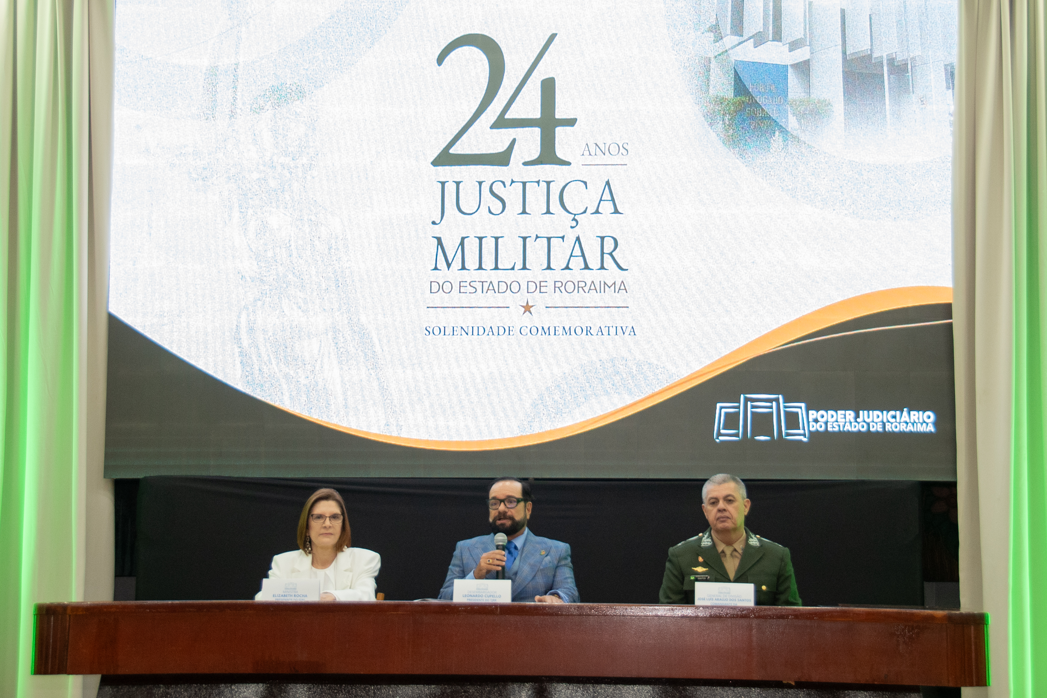 Imagem colorida registra a mesa de autoridades durante a solenidade comemorativa dos 24 anos da Justiça Militar de Roraima. Da esquerda para a direita, estão a presidente do Superior Tribunal Militar (STM), ministra Maria Elizabeth Guimarães Teixeira Rocha; o presidente do TJRR, desembargador Leonardo Cupello, falando ao microfone; e o general de Divisão José Luís Araújo dos Santos, comandante da Operação Acolhida. Ao fundo, painel exibe a identidade visual dos 24 anos da Justiça Militar do Estado de Roraima. Imagem colorida registra a mesa de autoridades durante a solenidade comemorativa dos 24 anos da Justiça Militar de Roraima. Da esquerda para a direita, estão a presidente do Superior Tribunal Militar (STM), ministra Maria Elizabeth Guimarães Teixeira Rocha; o presidente do TJRR, desembargador Leonardo Cupello, falando ao microfone; e o general de Divisão José Luís Araújo dos Santos, comandante da Operação Acolhida. Ao fundo, painel exibe a identidade visual dos 24 anos da Justiça Militar do Estado de Roraima.