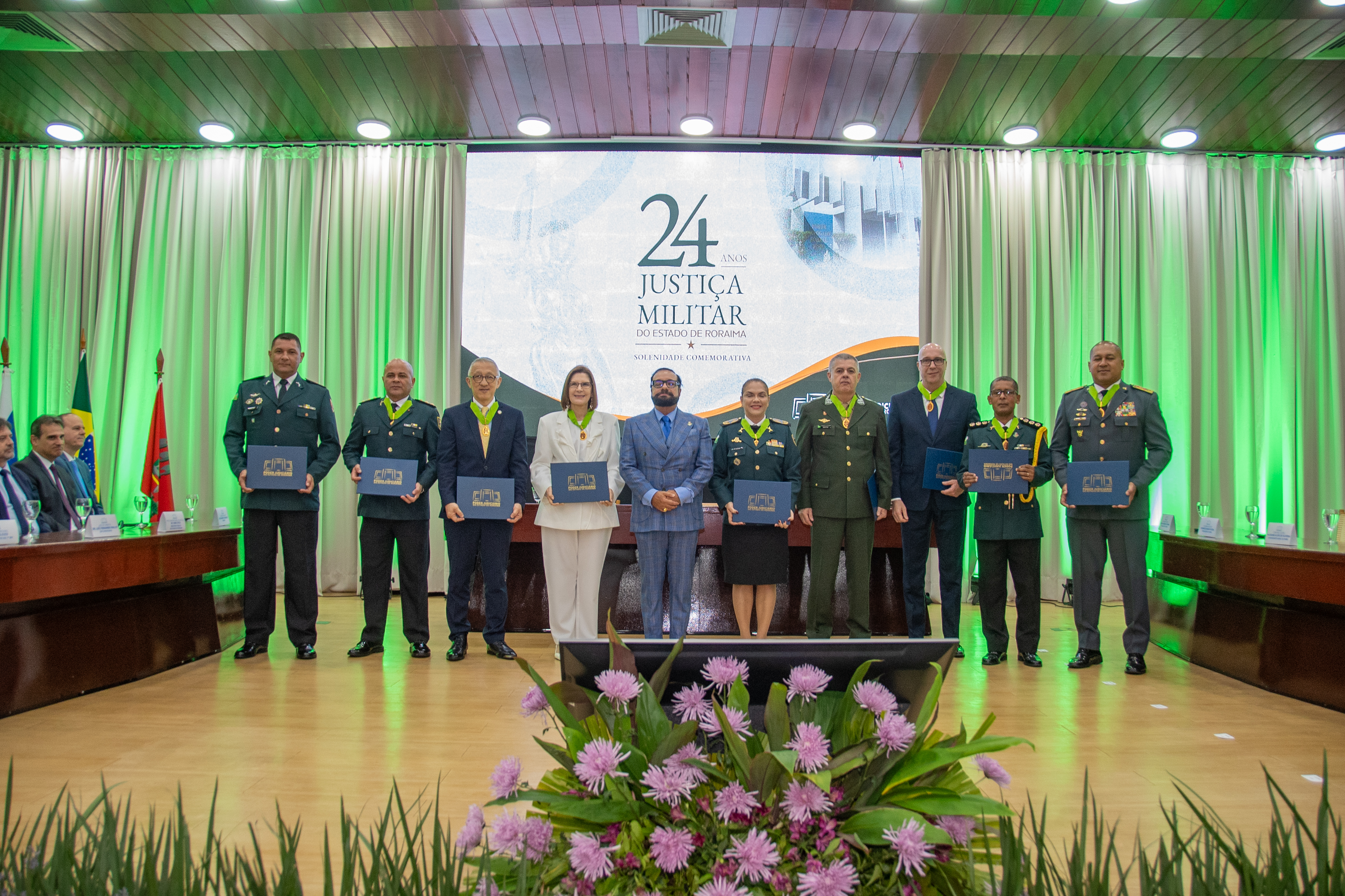 Imagem colorida registra os homenageados da solenidade comemorativa dos 24 anos da Justiça Militar de Roraima posando para fotografia oficial ao lado do presidente do TJRR, desembargador Leonardo Cupello. Militares e autoridades exibem diplomas e condecorações recebidos durante a cerimônia, diante do painel comemorativo do evento e da iluminação verde do auditório. Imagem colorida registra os homenageados da solenidade comemorativa dos 24 anos da Justiça Militar de Roraima posando para fotografia oficial ao lado do presidente do TJRR, desembargador Leonardo Cupello. Militares e autoridades exibem diplomas e condecorações recebidos durante a cerimônia, diante do painel comemorativo do evento e da iluminação verde do auditório.