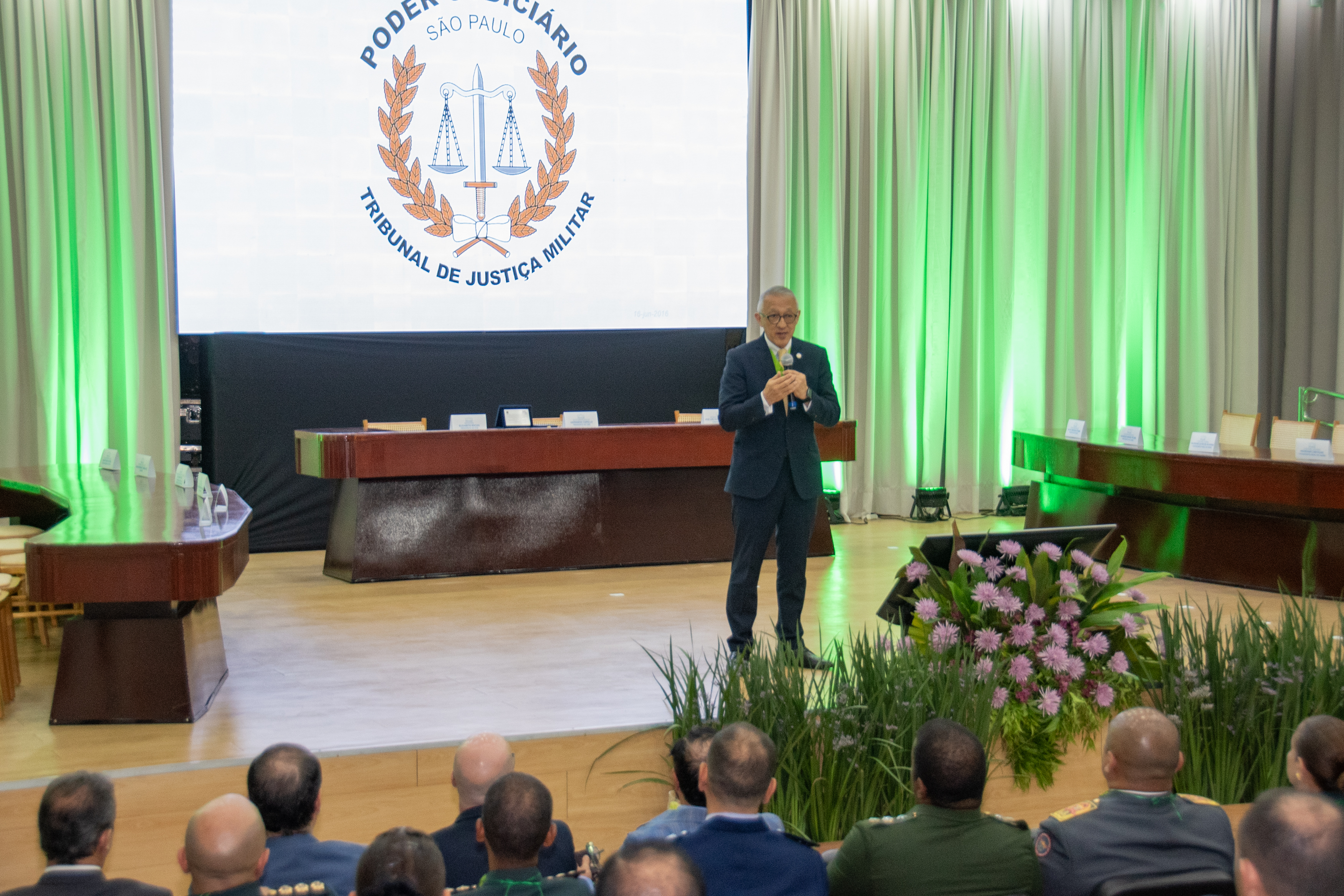 Imagem colorida registra o presidente do Tribunal de Justiça Militar de São Paulo (TJMSP), desembargador militar Silvio Hiroshi Oyama, durante palestra na solenidade comemorativa dos 24 anos da Justiça Militar de Roraima. Ele fala ao microfone diante do público, enquanto o painel ao fundo exibe o símbolo do Tribunal de Justiça Militar de São Paulo. Imagem colorida registra o presidente do Tribunal de Justiça Militar de São Paulo (TJMSP), desembargador militar Silvio Hiroshi Oyama, durante palestra na solenidade comemorativa dos 24 anos da Justiça Militar de Roraima. Ele fala ao microfone diante do público, enquanto o painel ao fundo exibe o símbolo do Tribunal de Justiça Militar de São Paulo.