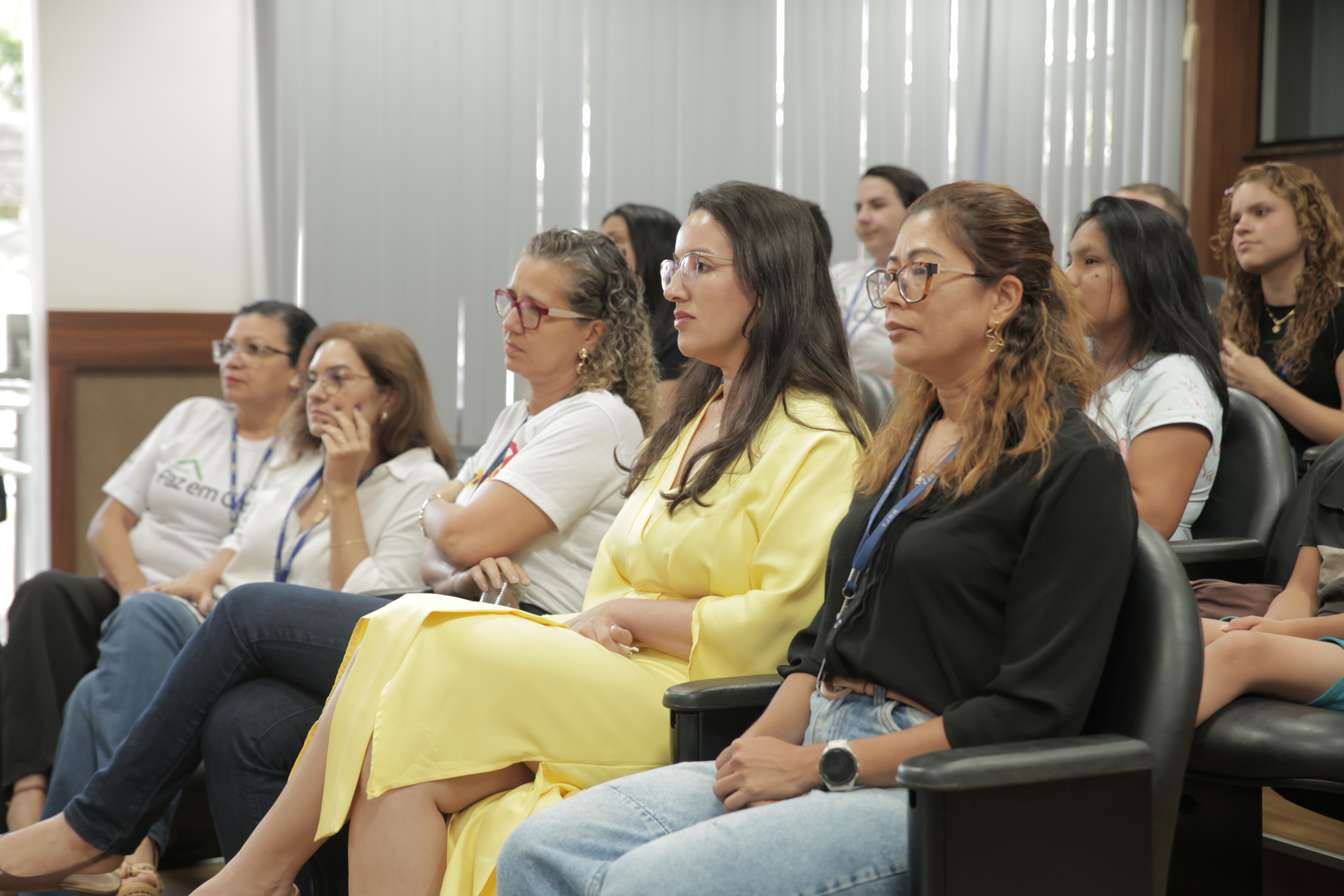 Foto 2: Fotografia colorida mostra um grupo de pessoas sentadas em poltronas pretas na plateia de um auditório, prestando atenção a um evento. Em destaque na primeira fileira, estão uma mulher de camisa preta e óculos e, ao lado dela, a palestrante de vestido amarelo. Foto 2: Fotografia colorida mostra um grupo de pessoas sentadas em poltronas pretas na plateia de um auditório, prestando atenção a um evento. Em destaque na primeira fileira, estão uma mulher de camisa preta e óculos e, ao lado dela, a palestrante de vestido amarelo.