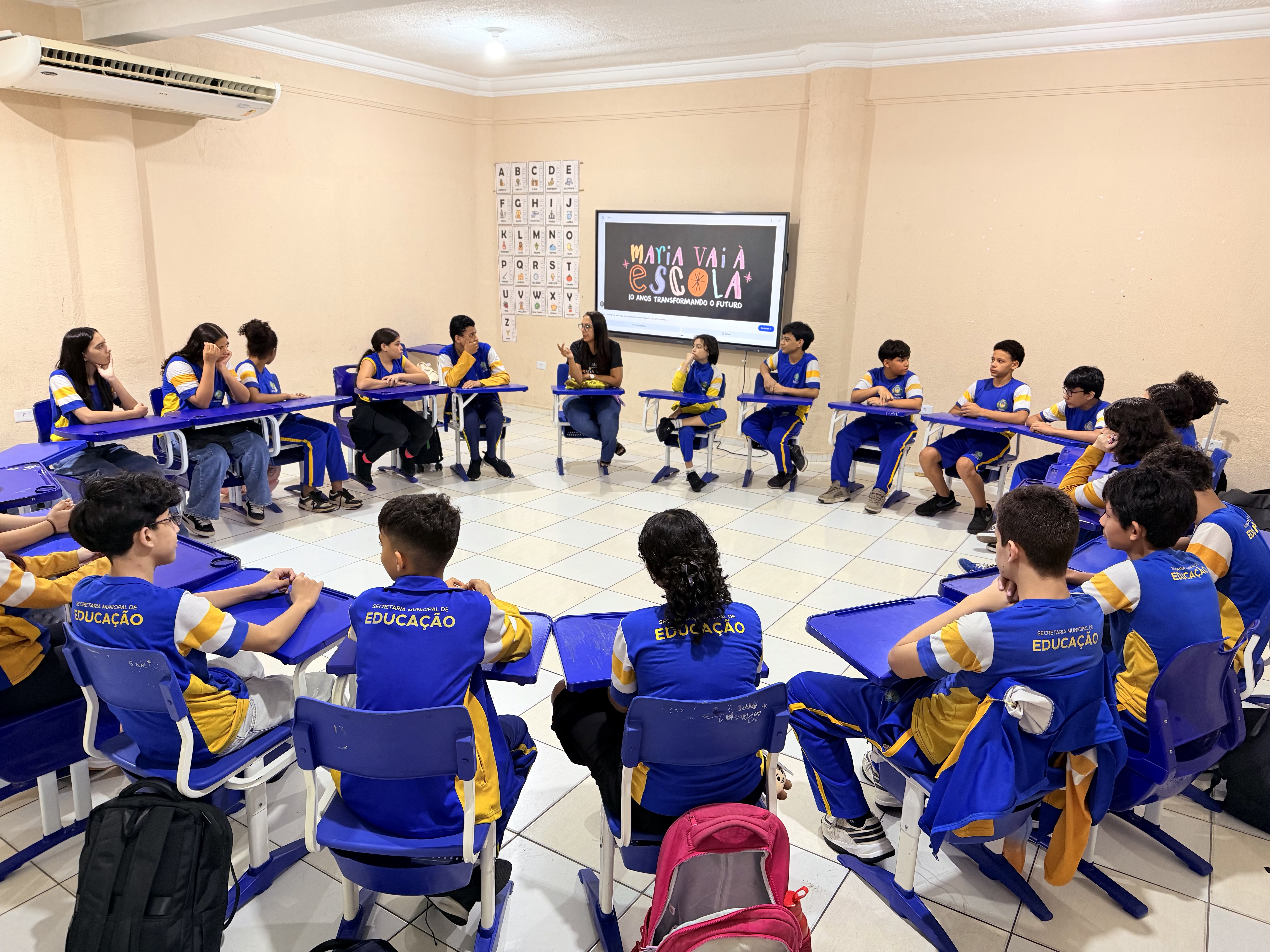 Imagem colorida mostra estudantes uniformizados e uma educadora dispostos em uma grande roda de carteiras em sala de aula. Ao fundo, uma tela exibe a apresentação do projeto "Maria Vai à Escola – 10 anos transformando o futuro".