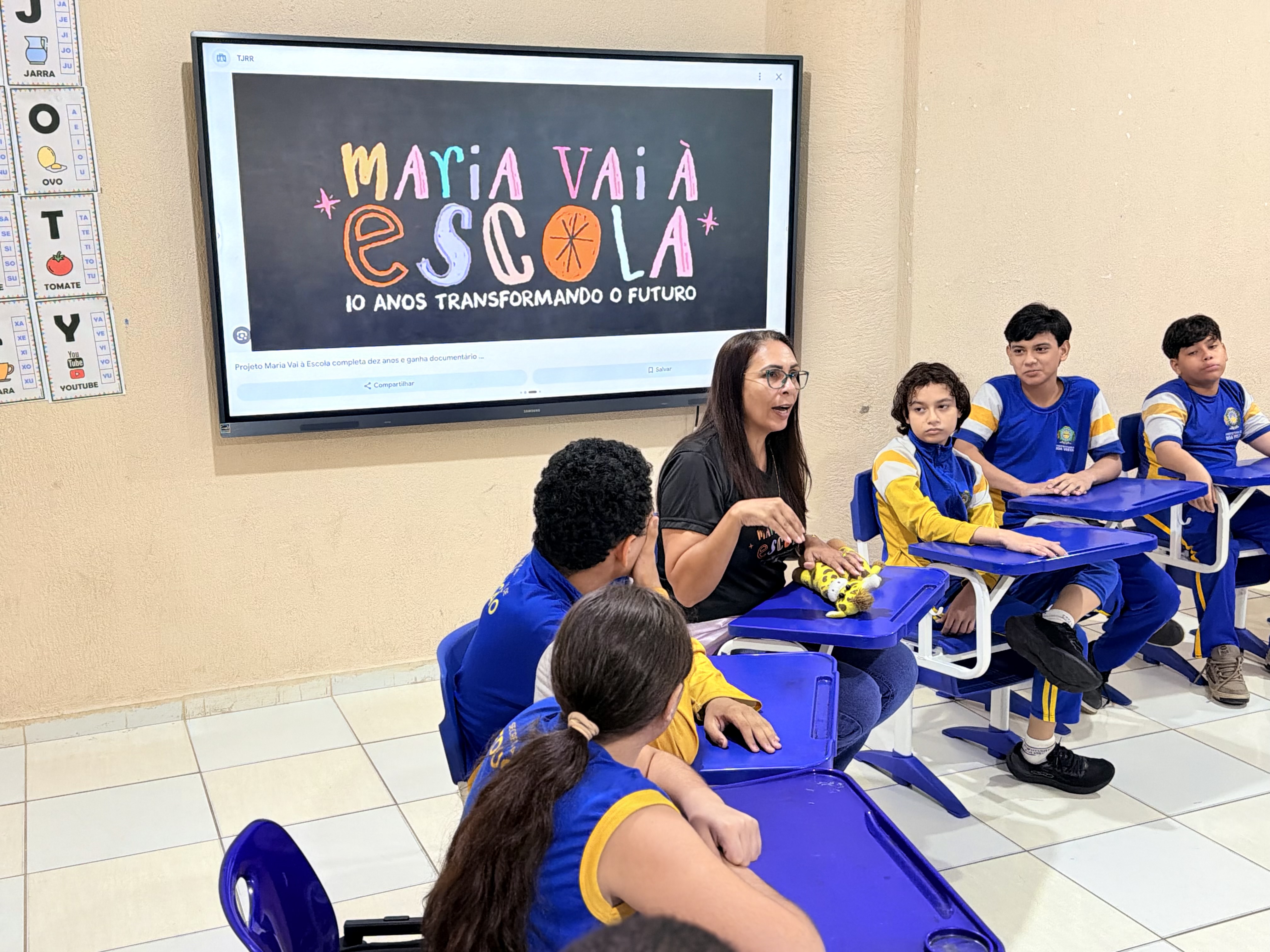 Imagem colorida mostra uma educadora conversando com estudantes em carteiras azuis durante atividade de sala de aula. Ao fundo, uma tela digital exibe o título da apresentação: "Maria Vai à Escola – 10 anos transformando o futuro".
