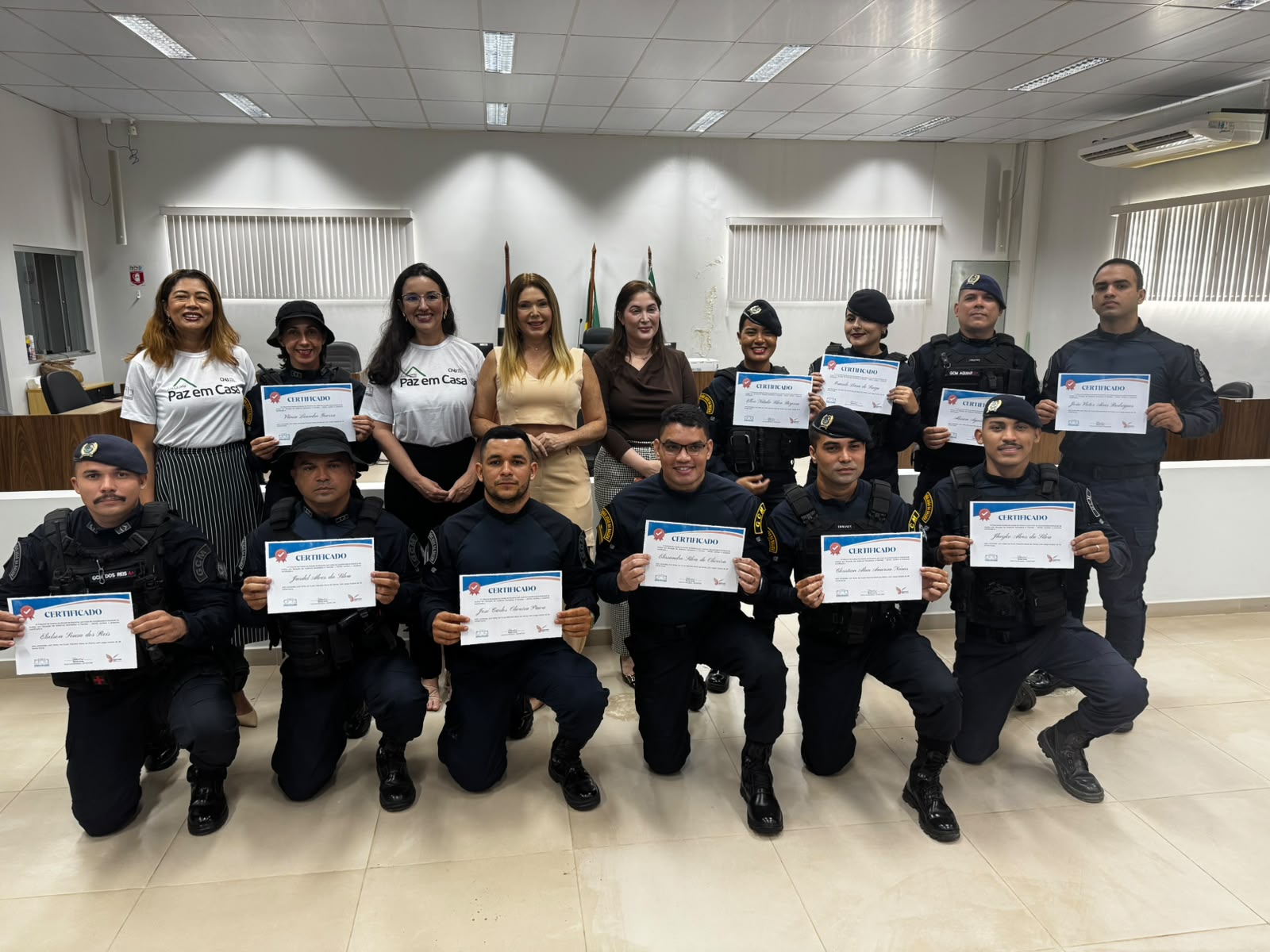 Imagem colorida mostra guardas municipais e organizadoras reunidos em uma sala para entrega de certificados. À frente, guardas ajoelhados exibem seus certificados, enquanto no fundo o restante do grupo permanece de pé com seus documentos.