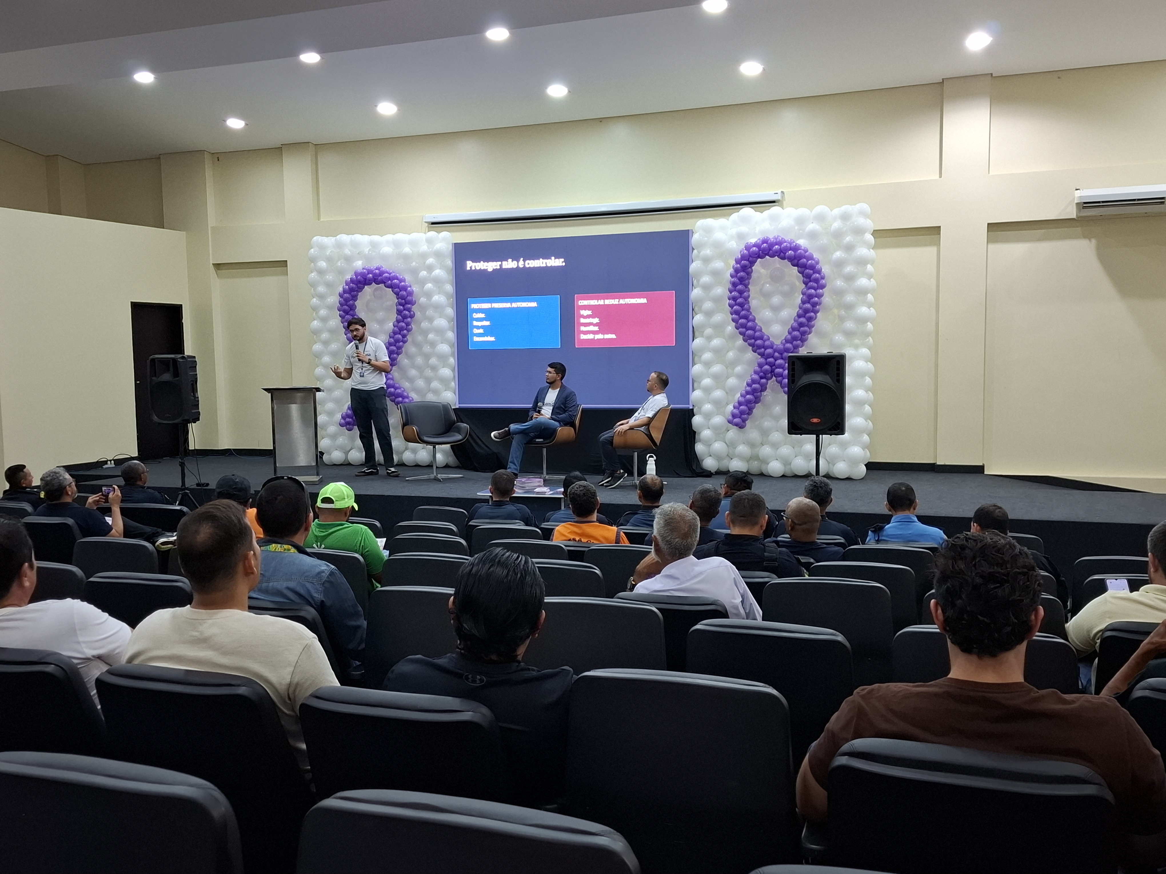  Imagem colorida mostra um palestrante de pé no palco apresentando uma palestra em um auditório lotado. No palco, decorado com painéis de balões brancos e roxos no formato do laço do Agosto Lilás, dois homens estão sentados em poltronas enquanto a plateia assiste de costas em primeiro plano.