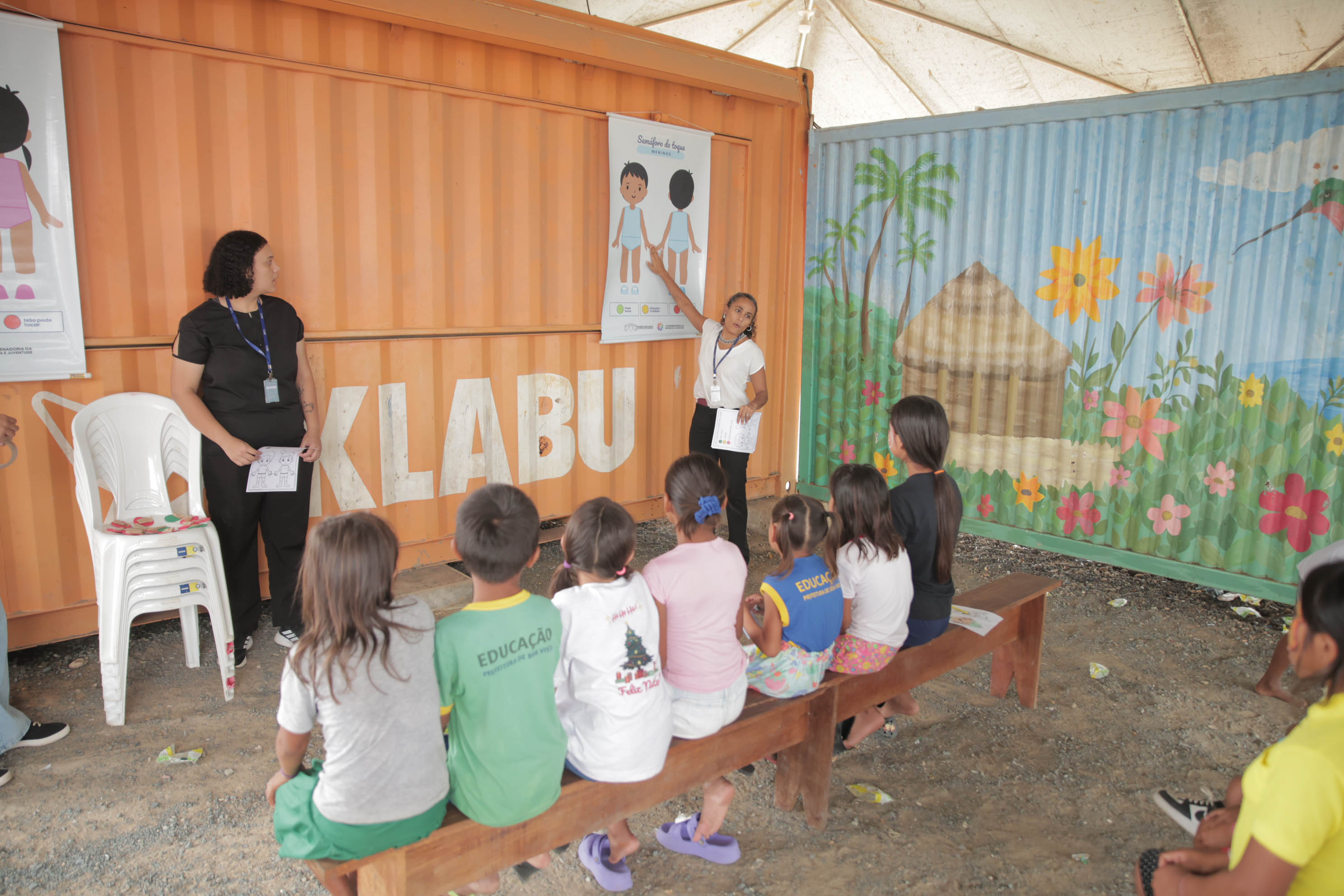 Fotografia de uma atividade educativa da CIJ para crianças sentadas em um banco de madeira, acompanhando a explicação do cartaz "Semáforo do toque" afixado em um container. Fotografia de uma atividade educativa da CIJ para crianças sentadas em um banco de madeira, acompanhando a explicação do cartaz "Semáforo do toque" afixado em um container.