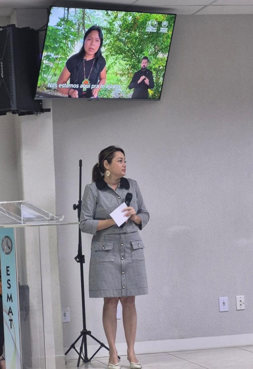 Fotografia de uma palestrante em pé no palco segurando um microfone e um papel. Acima dela, um monitor exibe um vídeo com uma mulher indígena e um intérprete de Libras. À esquerda, destaca-se um púlpito com a sigla ESMAT.