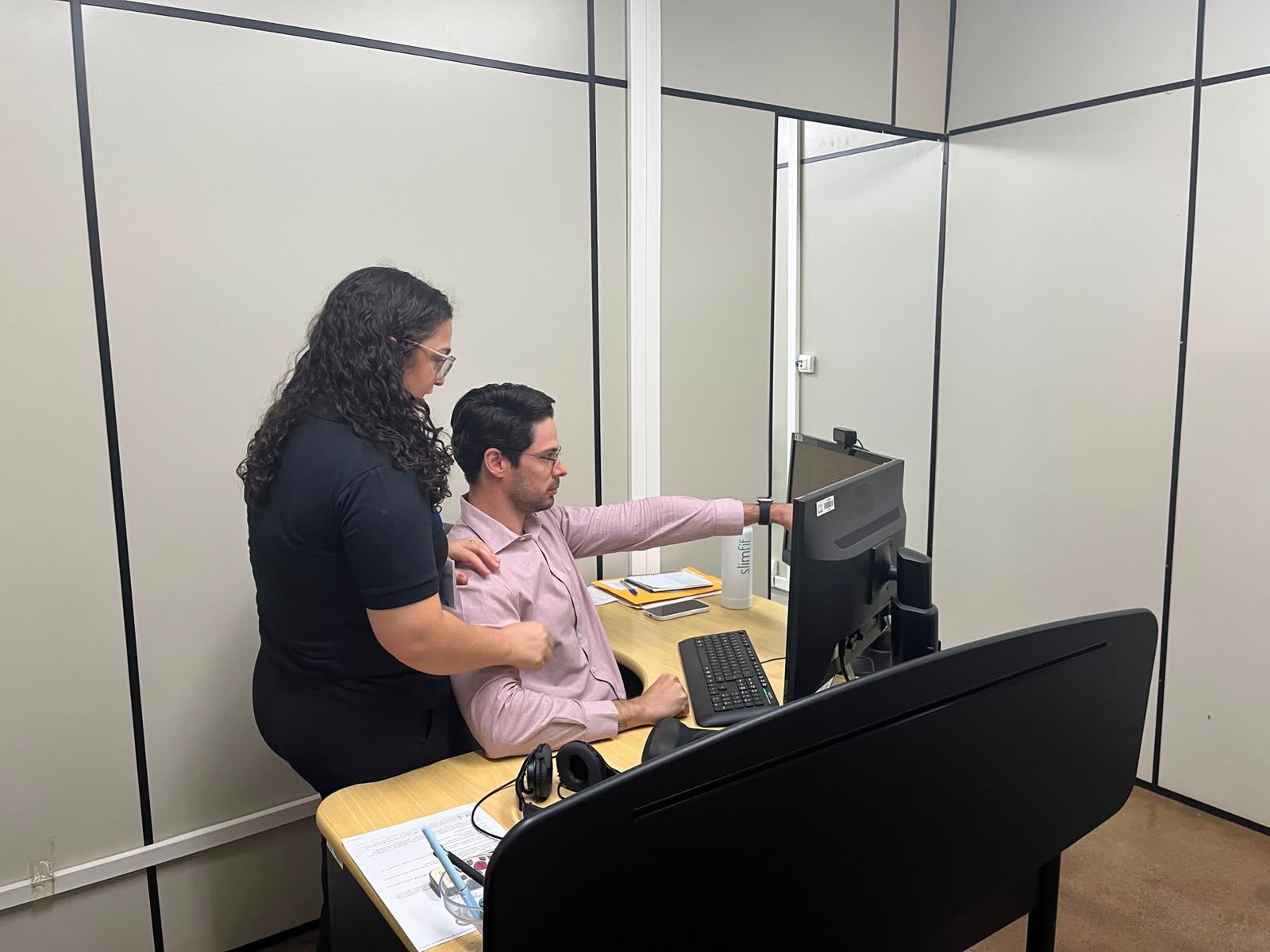 Imagem colorida registra uma orientação ergonômica em uma estação de trabalho. Um homem permanece sentado em frente ao computador e aponta para a tela, enquanto uma mulher acompanha o posicionamento e orienta a postura e a altura adequada da cadeira. Imagem colorida registra uma orientação ergonômica em uma estação de trabalho. Um homem permanece sentado em frente ao computador e aponta para a tela, enquanto uma mulher acompanha o posicionamento e orienta a postura e a altura adequada da cadeira.