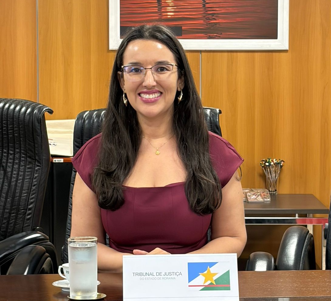 Imagem colorida registra uma mulher sentada à mesa durante atividade institucional. Ela sorri para a câmera diante de uma placa de identificação do Tribunal de Justiça do Estado de Roraima, em uma sala de reunião. Imagem colorida registra uma mulher sentada à mesa durante atividade institucional. Ela sorri para a câmera diante de uma placa de identificação do Tribunal de Justiça do Estado de Roraima, em uma sala de reunião.