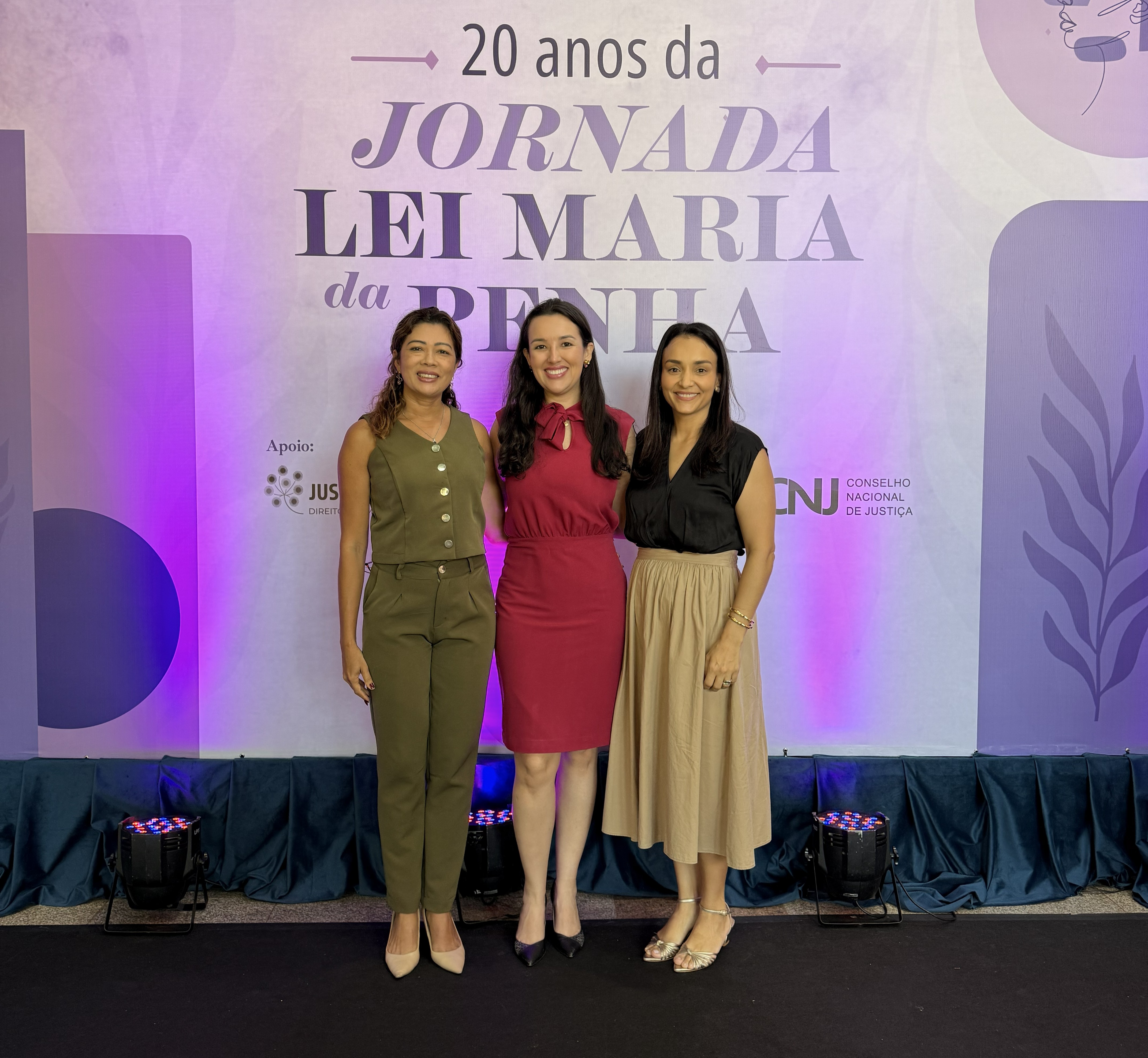 Imagem colorida mostra três mulheres posando para fotografia durante a XX Jornada da Lei Maria da Penha. Elas aparecem sorrindo diante do painel comemorativo dos 20 anos da Jornada, com a identificação do Conselho Nacional de Justiça ao fundo. Imagem colorida mostra três mulheres posando para fotografia durante a XX Jornada da Lei Maria da Penha. Elas aparecem sorrindo diante do painel comemorativo dos 20 anos da Jornada, com a identificação do Conselho Nacional de Justiça ao fundo.