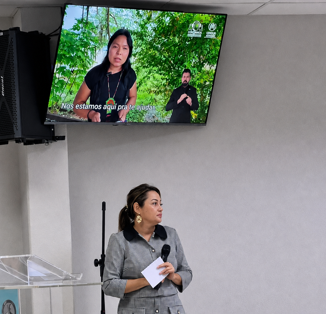 Fotografia de uma palestrante em pé no palco segurando um microfone e um papel. Acima dela, um monitor exibe um vídeo com uma mulher indígena e um intérprete de Libras. À esquerda, destaca-se um púlpito com a sigla ESMAT. Fotografia de uma palestrante em pé no palco segurando um microfone e um papel. Acima dela, um monitor exibe um vídeo com uma mulher indígena e um intérprete de Libras. À esquerda, destaca-se um púlpito com a sigla ESMAT.