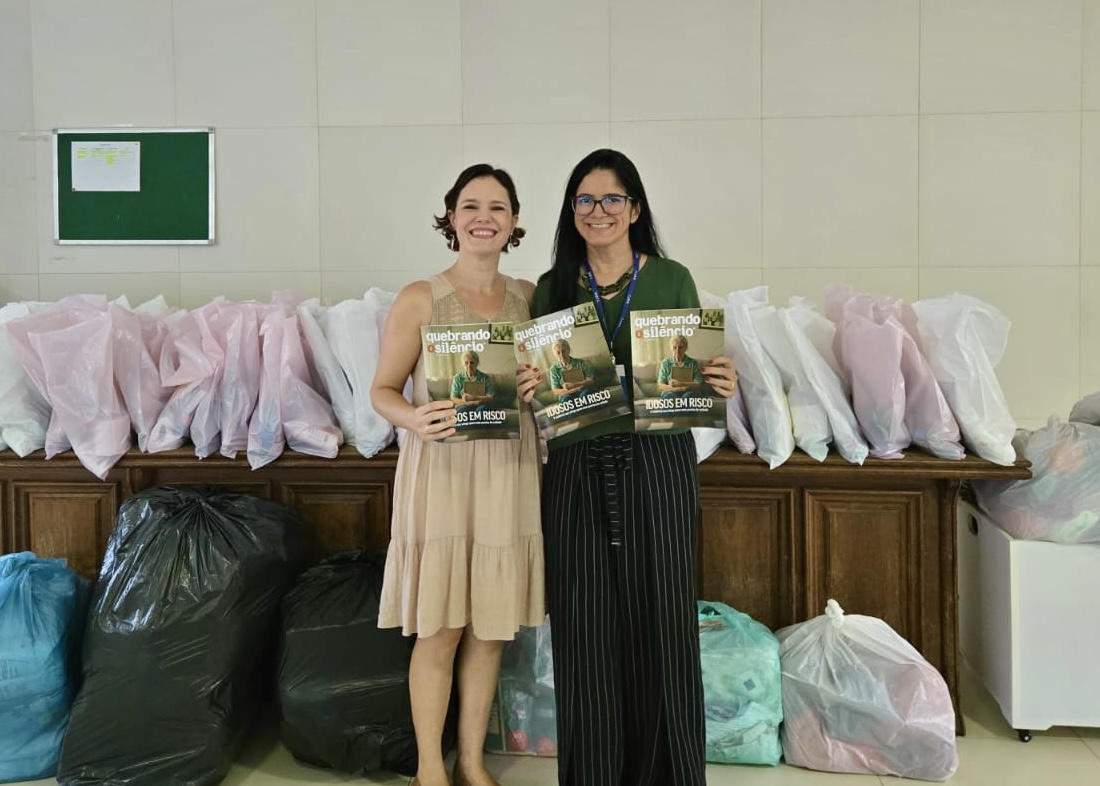 Foto 2: Imagem colorida mostra duas mulheres sorrindo enquanto exibem exemplares da revista "Quebrando o Silêncio: Idosos em Risco". Elas estão posicionadas lado a lado em frente a balcões e sacolas de doações organizadas no ambiente. Foto 2: Imagem colorida mostra duas mulheres sorrindo enquanto exibem exemplares da revista "Quebrando o Silêncio: Idosos em Risco". Elas estão posicionadas lado a lado em frente a balcões e sacolas de doações organizadas no ambiente.
