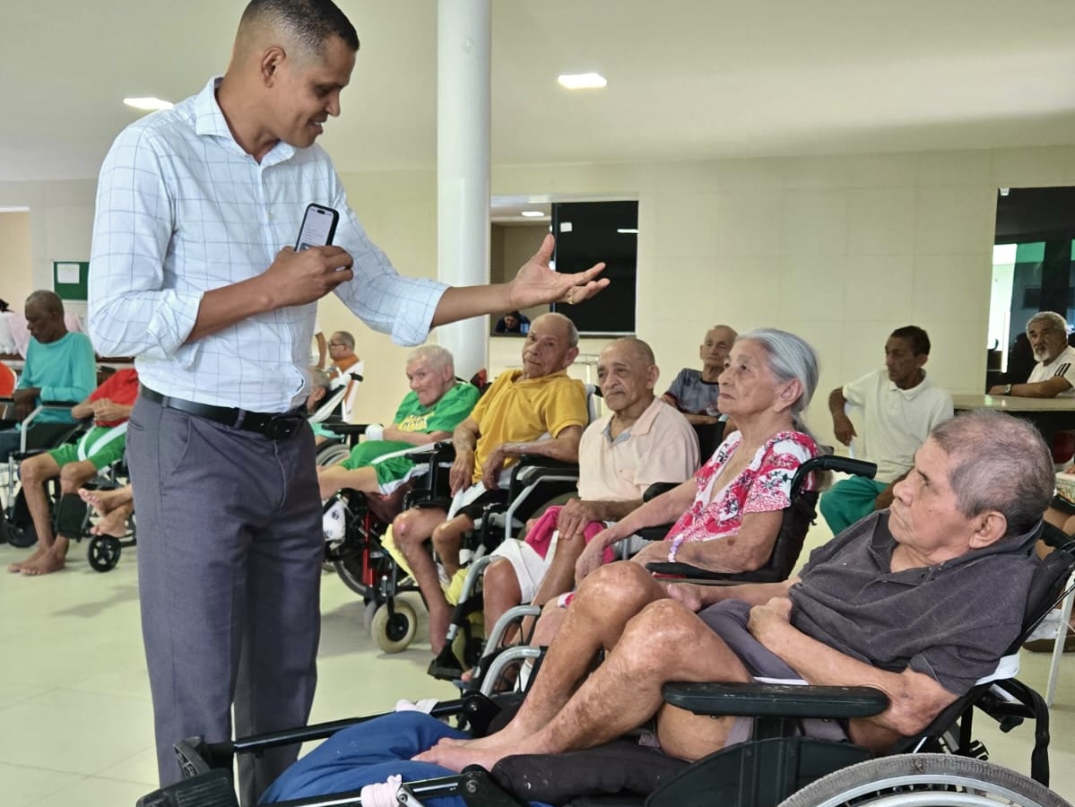 Foto 3: Imagem colorida mostra um homem de camisa social quadriculada clara e calça social de pé, sorrindo e gesticulando enquanto conversa com idosos em um salão. À sua frente, idosos acomodados em cadeiras de rodas e cadeiras comuns escutam atentamente a interação. Foto 3: Imagem colorida mostra um homem de camisa social quadriculada clara e calça social de pé, sorrindo e gesticulando enquanto conversa com idosos em um salão. À sua frente, idosos acomodados em cadeiras de rodas e cadeiras comuns escutam atentamente a interação.