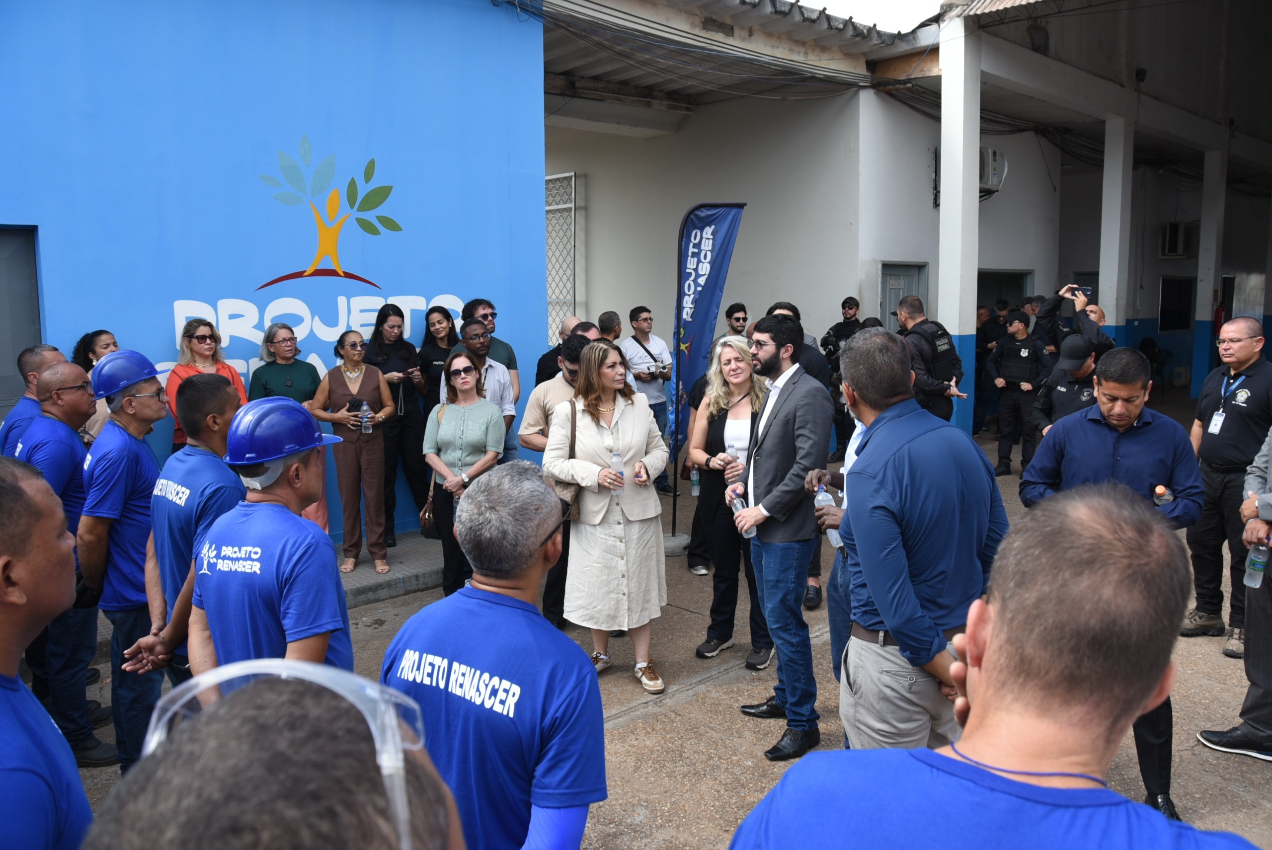 Fotografia mostra um grupo de pessoas reunidas em frente a um prédio. Em primeiro plano, trabalhadores vestindo camisetas azuis com a identificação “Projeto Renascer” estão de costas para a câmera. Ao centro, outras pessoas acompanham a atividade. Ao fundo, há uma parede azul com a identidade visual do Projeto Renascer e uma faixa vertical com o nome do projeto.
