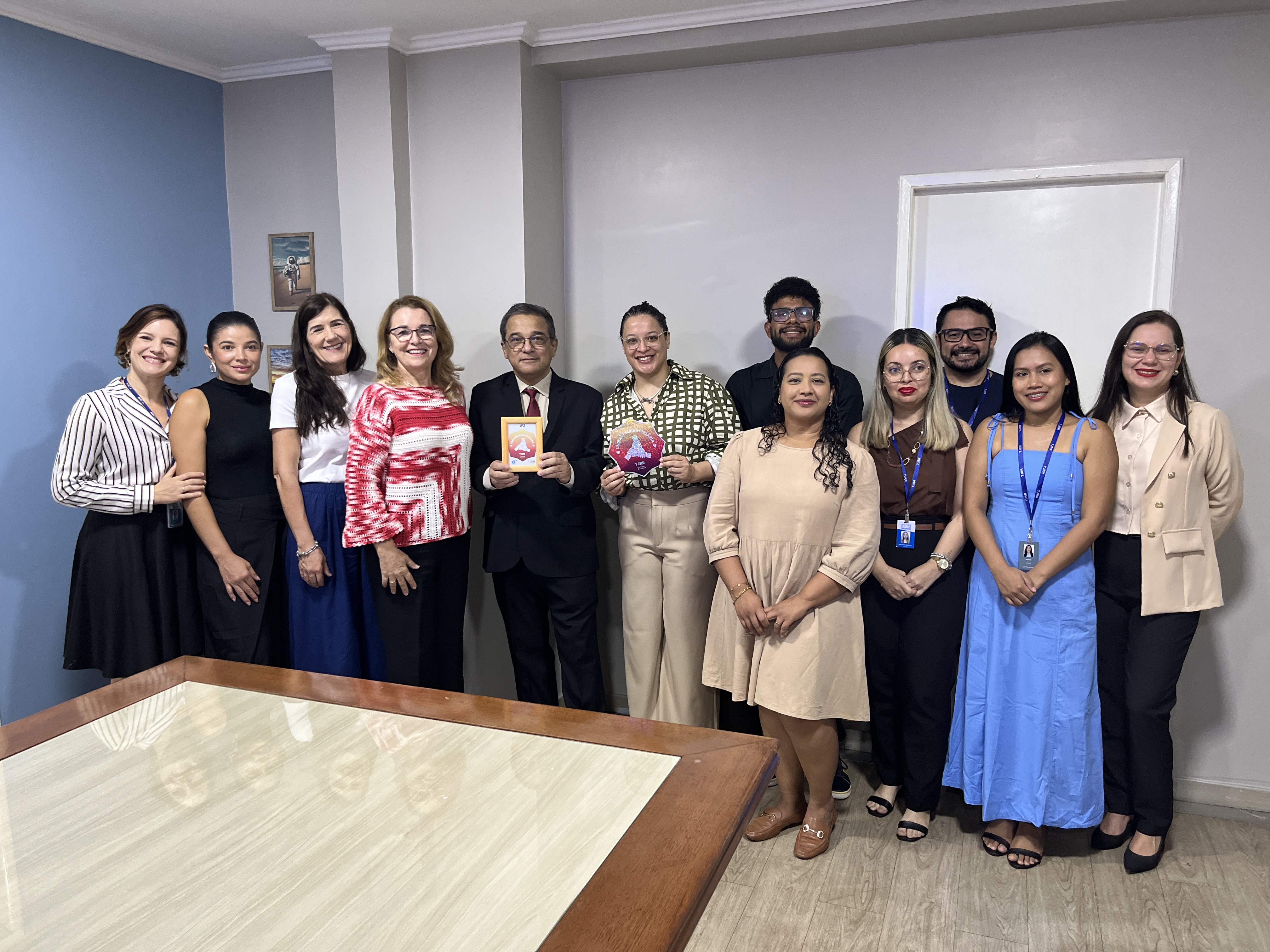 Imagem colorida mostra um grupo de onze pessoas posando sorridentes para uma foto na sala de reuniões. O homem de terno, posicionado ao centro, segura um pequeno quadro emoldurado, enquanto a mulher ao seu lado exibe a placa hexagonal colorida. Imagem colorida mostra um grupo de onze pessoas posando sorridentes para uma foto na sala de reuniões. O homem de terno, posicionado ao centro, segura um pequeno quadro emoldurado, enquanto a mulher ao seu lado exibe a placa hexagonal colorida.