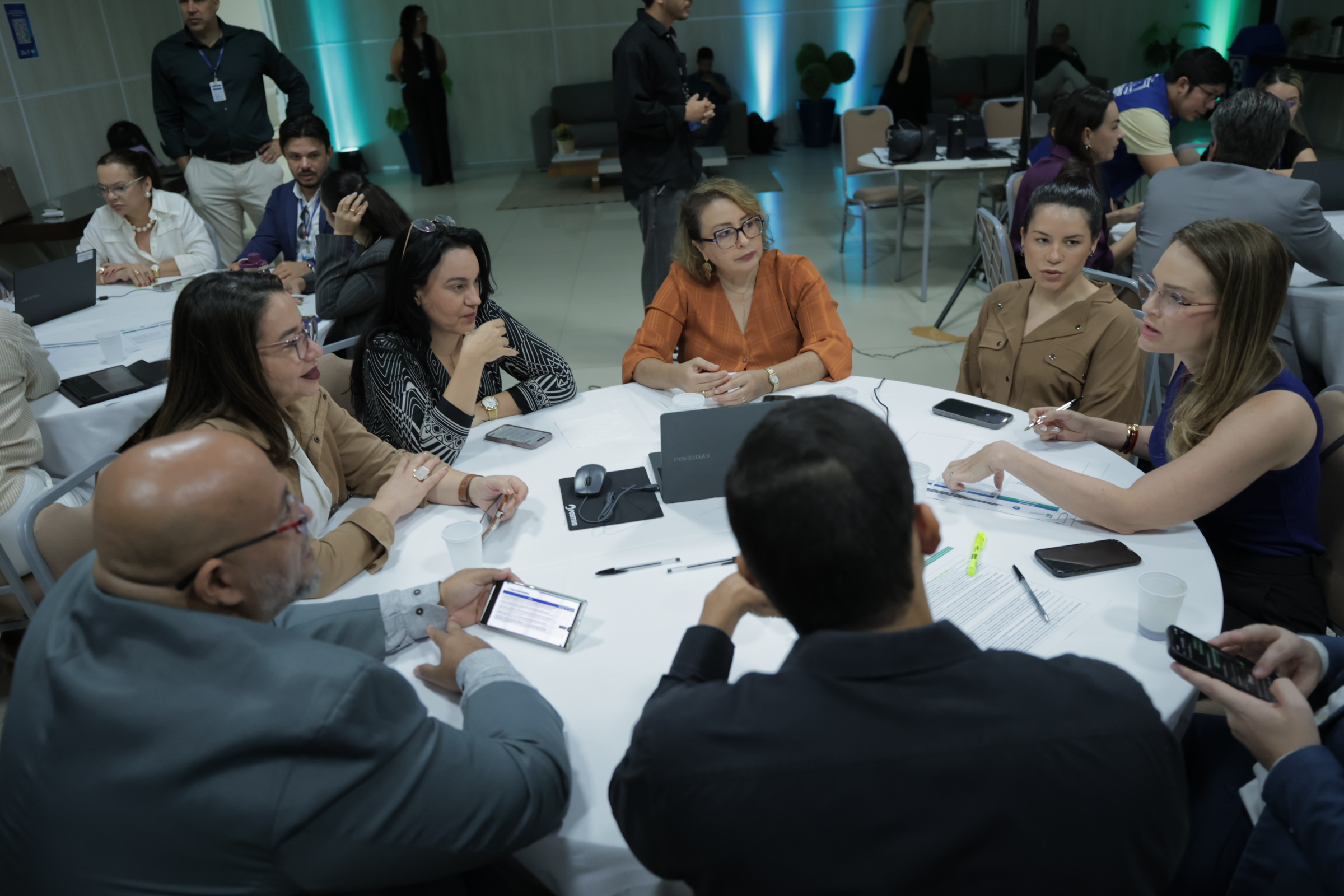 Fotografia mostra um grupo de pessoas reunido ao redor de uma mesa durante uma atividade de trabalho. Os participantes conversam e analisam documentos, utilizando computadores, tablets, celulares e outros materiais. Ao fundo, há outras mesas com participantes. Fotografia mostra um grupo de pessoas reunido ao redor de uma mesa durante uma atividade de trabalho. Os participantes conversam e analisam documentos, utilizando computadores, tablets, celulares e outros materiais. Ao fundo, há outras mesas com participantes.