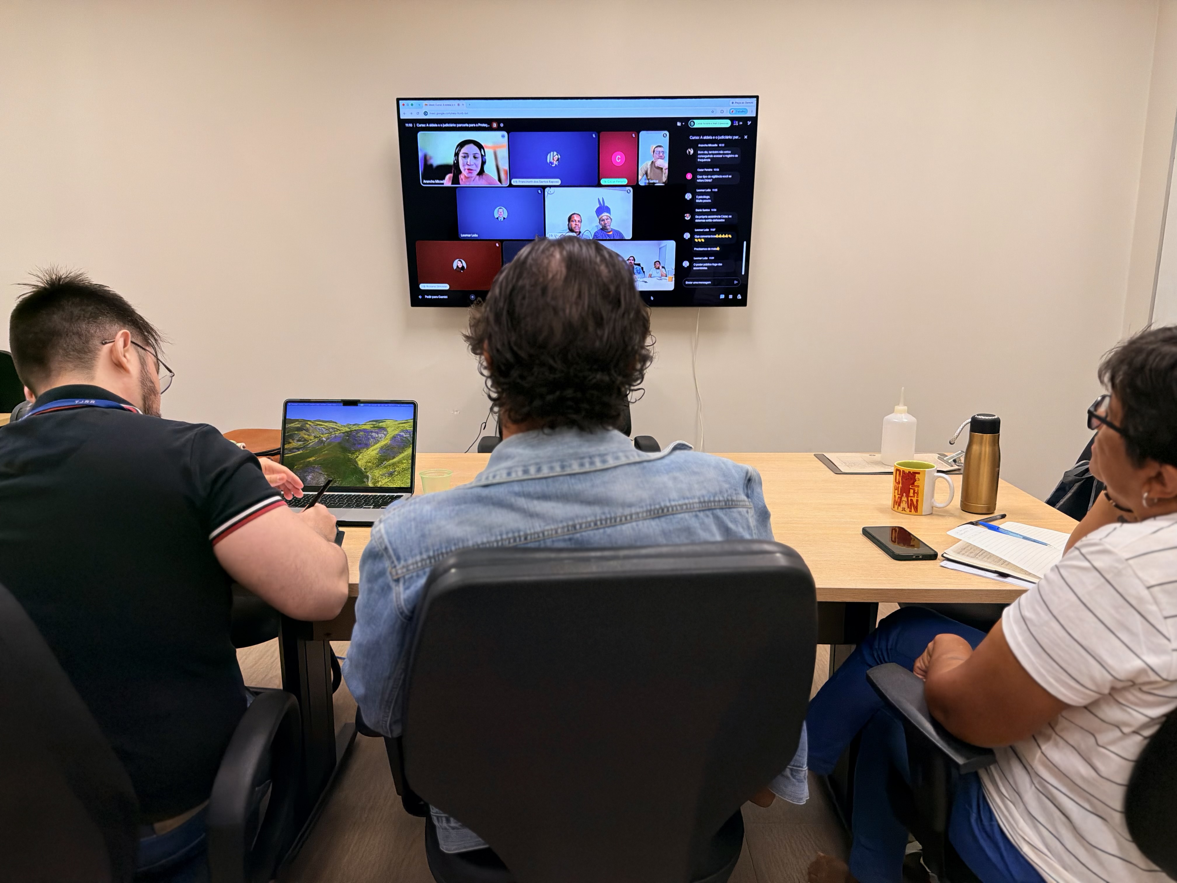 Imagem colorida mostra três pessoas de costas, dispostas em torno de uma mesa clara de reunião, observando uma televisão fixada na parede à frente. Na tela da TV, é exibida uma videochamada ao vivo no Google Meet com múltiplos participantes.