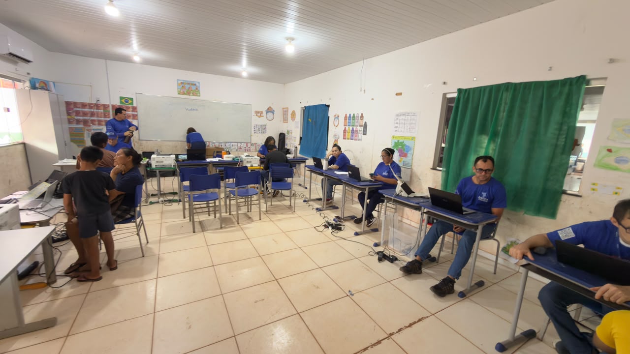 Imagem colorida mostra uma sala de aula adaptada para atendimento comunitário, com funcionários vestindo camisetas azuis e usando notebooks e impressoras em carteiras escolares dispostas pelo espaço enquanto atendem moradores locais sentados ao lado.