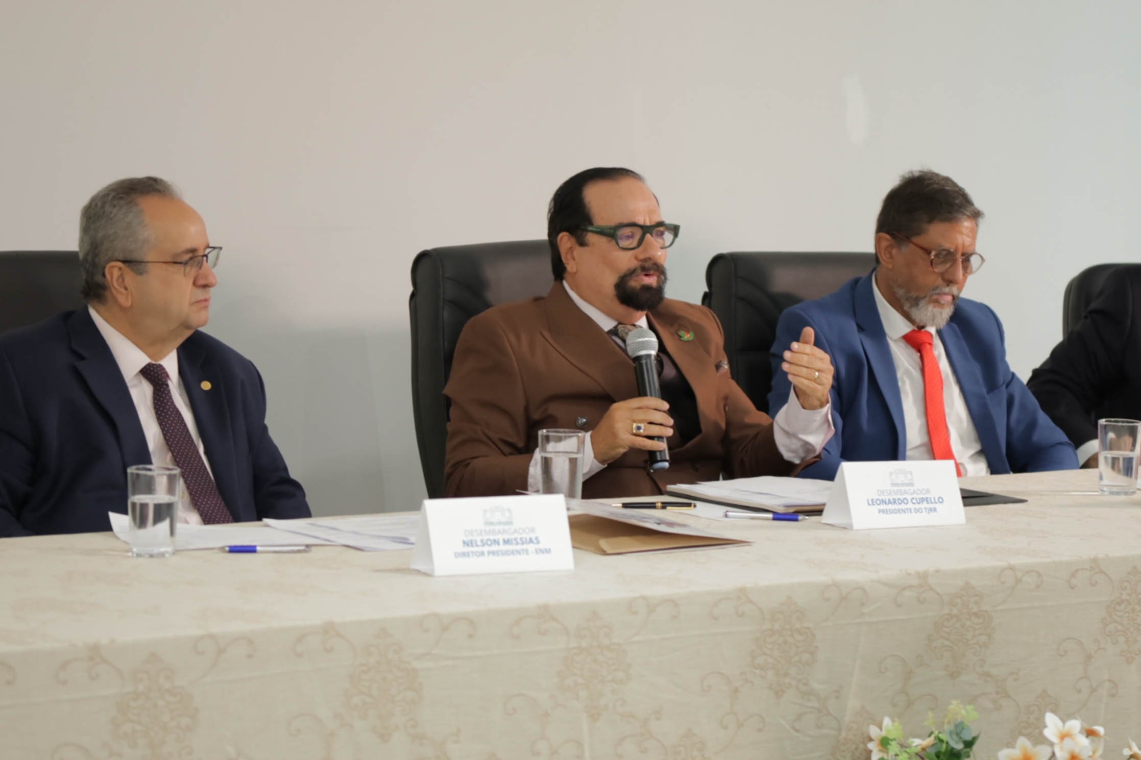 Imagem colorida registra o presidente do TJRR, desembargador Leonardo Cupello, durante o seminário sobre o método APAC, acompanhado por outros dois integrantes da mesa, com documentos e copos de água sobre a bancada.