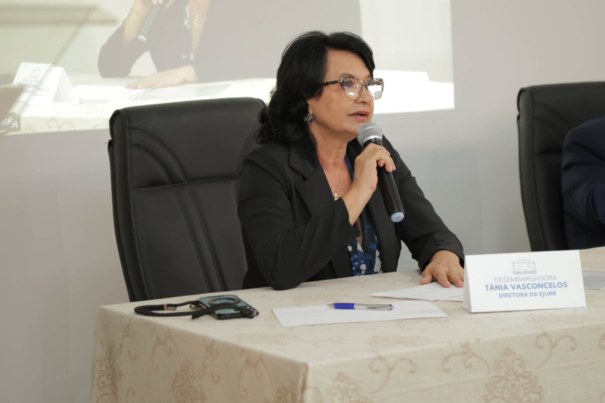 Imagem colorida mostra a diretora da EJURR, desembargadora Tânia Vasconcelos, falando ao microfone durante a programação do seminário. Sentada à mesa, ela aparece diante de documentos e de uma placa de identificação, enquanto acompanha as discussões sobre a metodologia APAC.