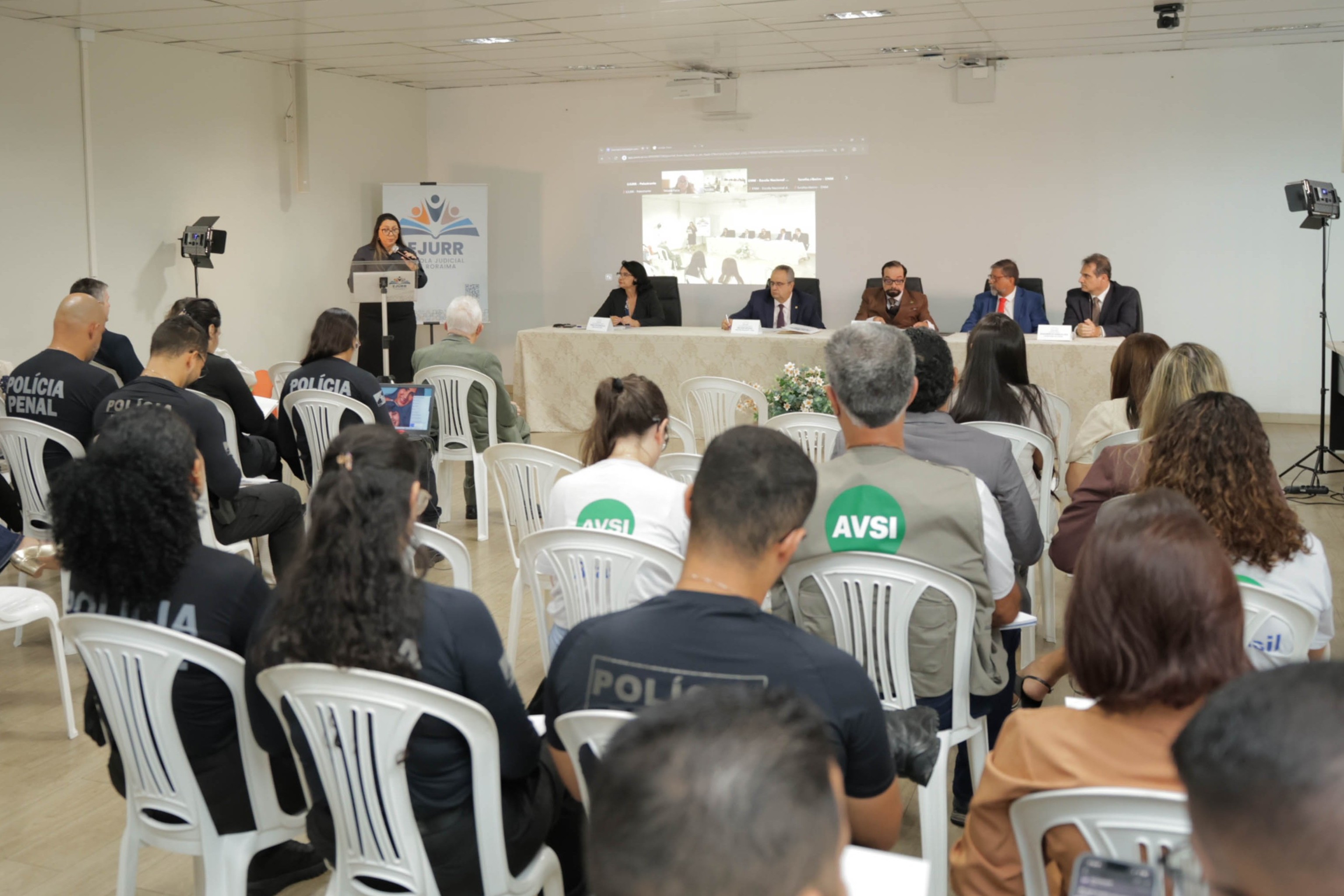 Imagem colorida apresenta visão ampla do seminário sobre o método APAC, com profissionais do Judiciário, do sistema prisional e de instituições parceiras acompanhando a programação. À frente, autoridades compõem a mesa e uma palestrante fala ao púlpito, enquanto uma videoconferência é projetada ao fundo.