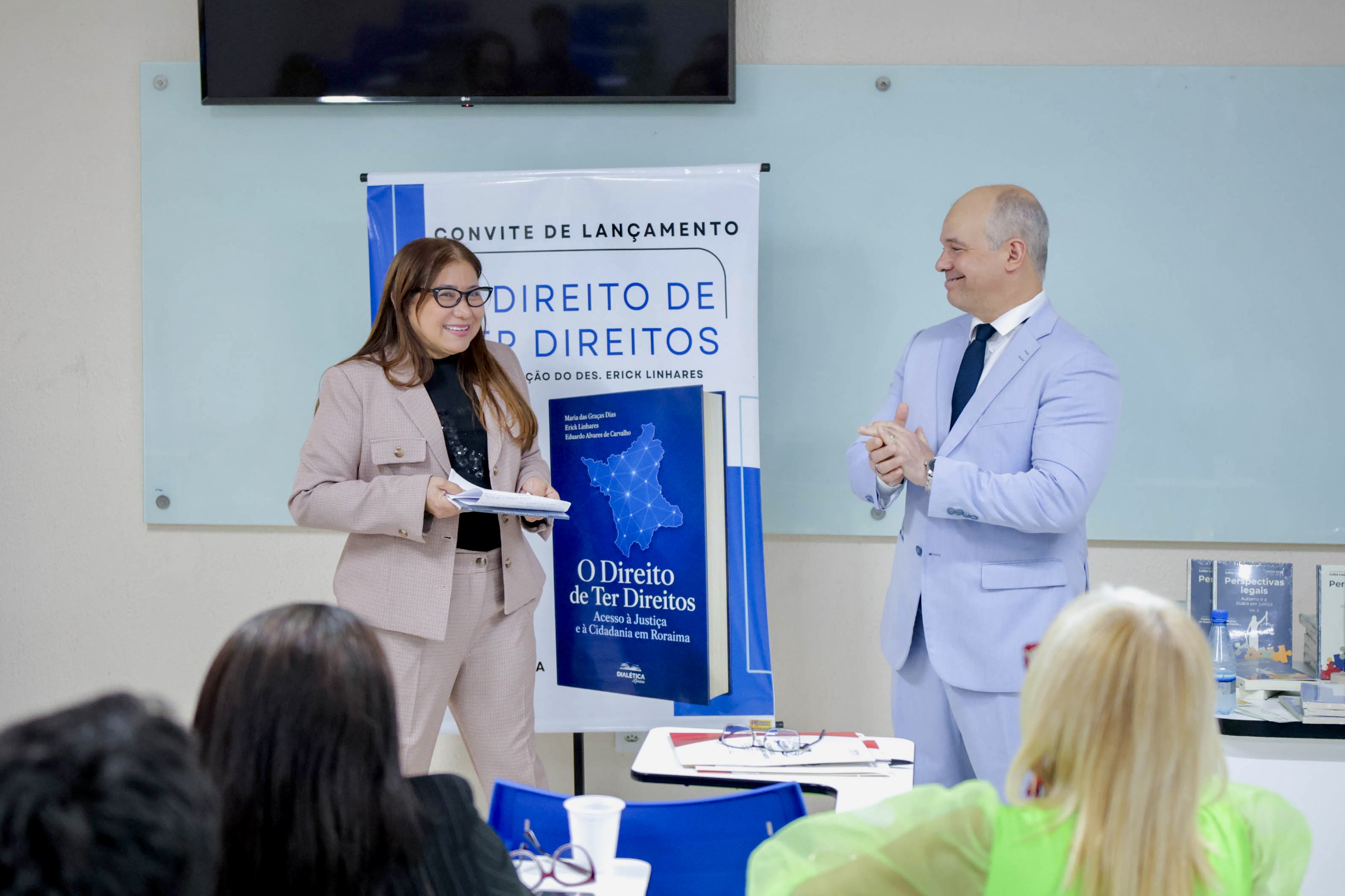Foto 2: Imagem colorida mostra evento de lançamento do livro "O Direito de Ter Direitos". Ao centro, em pé ao lado de um banner com a capa da obra, uma mulher de terninho bege segura um exemplar aberto enquanto sorri para um homem em terninho azul-claro que aplaude a ação. Em primeiro plano, parte do público assiste à apresentação. Fim da descrição. Foto 2: Imagem colorida mostra evento de lançamento do livro "O Direito de Ter Direitos". Ao centro, em pé ao lado de um banner com a capa da obra, uma mulher de terninho bege segura um exemplar aberto enquanto sorri para um homem em terninho azul-claro que aplaude a ação. Em primeiro plano, parte do público assiste à apresentação. Fim da descrição.