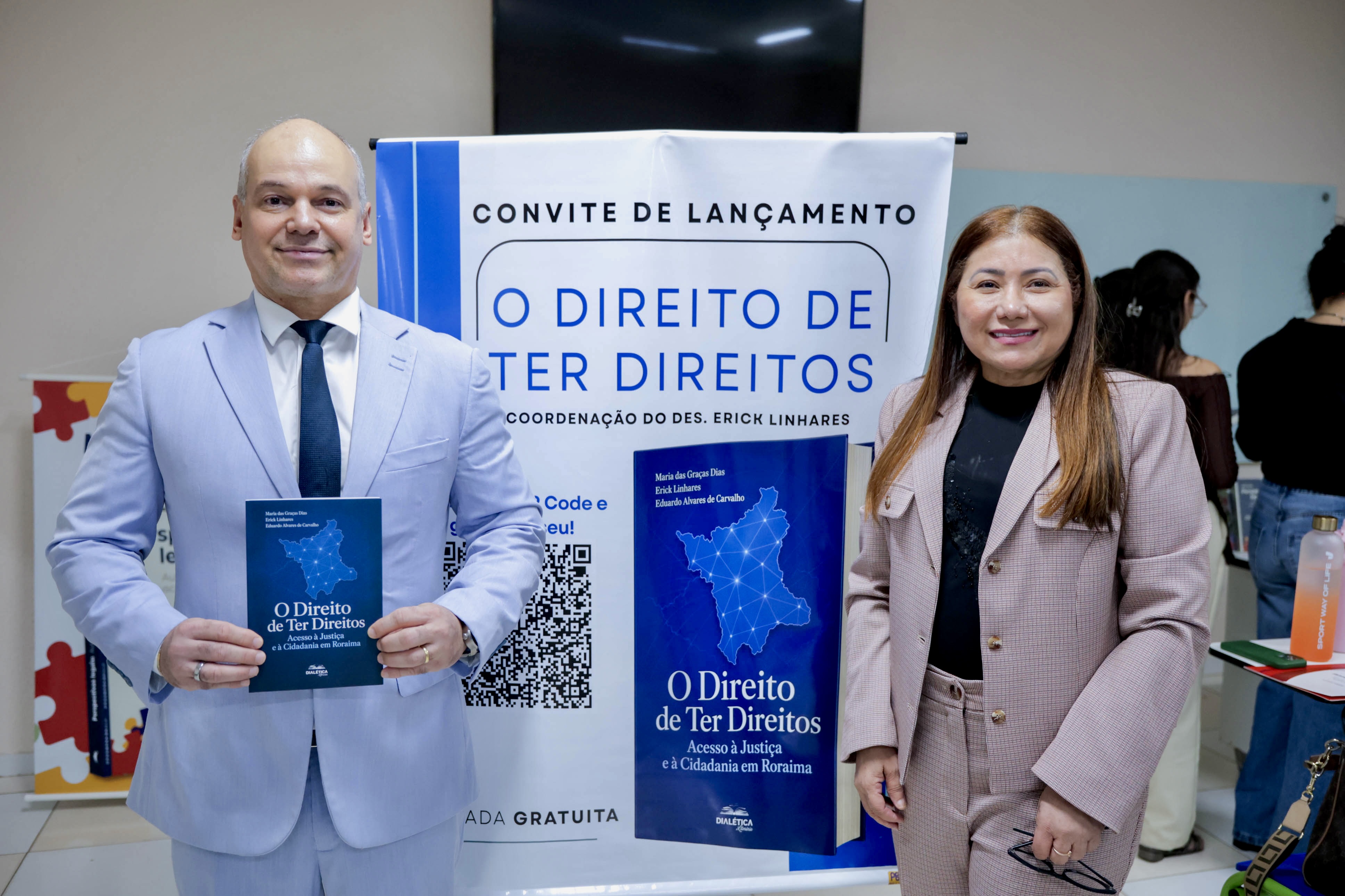 Foto 3: Imagem colorida colorida mostra um homem em terninho azul-claro segurando o livro "O Direito de Ter Direitos" ao lado de uma mulher em terninho bege. Ambos estão em pé à frente do banner do evento de lançamento do livro. Fim da descrição. Foto 3: Imagem colorida colorida mostra um homem em terninho azul-claro segurando o livro "O Direito de Ter Direitos" ao lado de uma mulher em terninho bege. Ambos estão em pé à frente do banner do evento de lançamento do livro. Fim da descrição.