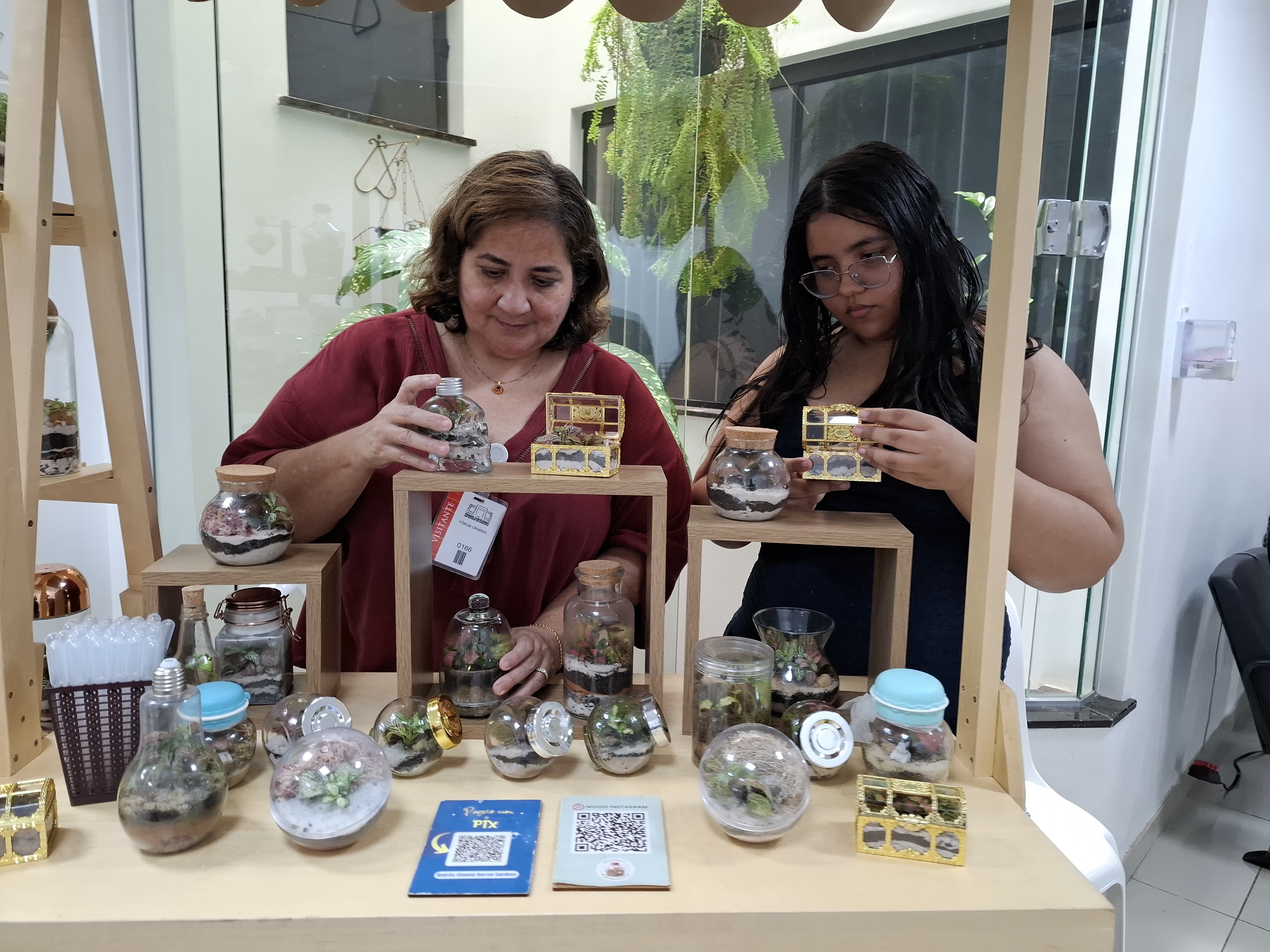 Imagem colorida mostra duas mulheres atrás de um pequeno estande de madeira organizando seus produtos. A bancada exibe diversos pequenos terrários de plantas em potes de vidro e caixinhas decorativas. Imagem colorida mostra duas mulheres atrás de um pequeno estande de madeira organizando seus produtos. A bancada exibe diversos pequenos terrários de plantas em potes de vidro e caixinhas decorativas.