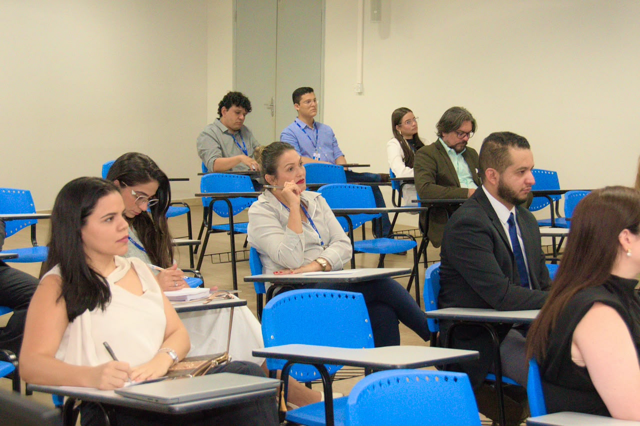Foto 2: Imagem colorida mostra participantes sentados em carteiras universitárias azuis assistindo atentamente a uma capacitação em sala de aula. Em primeiro plano, homens e mulheres vestindo trajes sociais e de passeio acompanham a apresentação e fazem anotações. Fim da descrição.