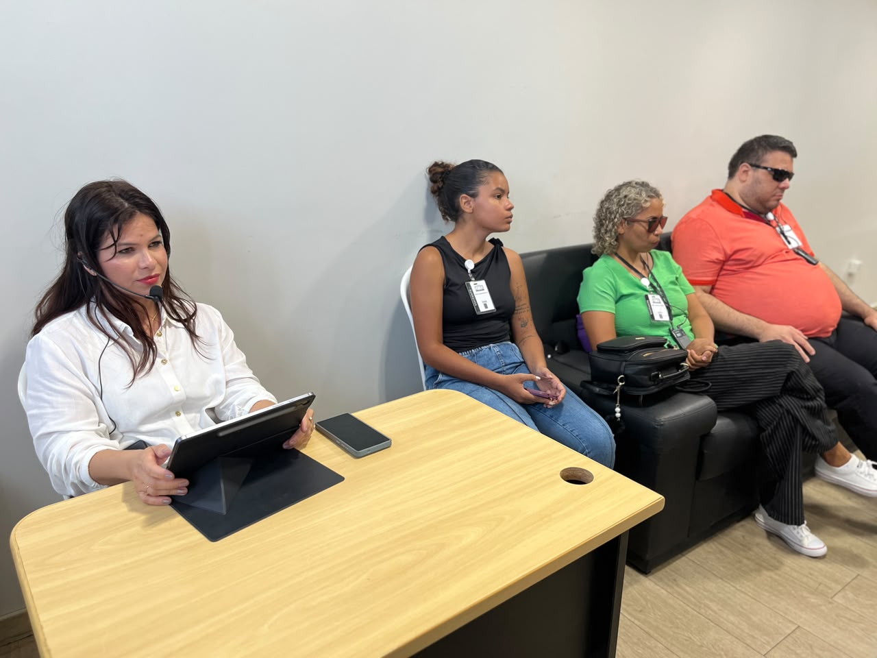 Fotografia de um serviço de acessibilidade em um evento onde uma profissional realiza audiodescrição em tempo real utilizando um fone de ouvido, tendo ao fundo participantes com deficiência visual sentados em um sofá acompanhando a transmissão e, em primeiro plano, a audiodescritora operando um tablet sobre uma mesa de madeira.