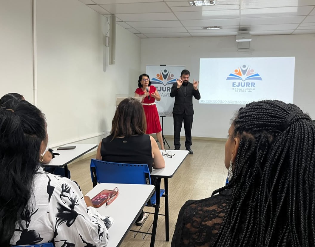 Fotografia de uma palestra com interpretação em Libras onde uma mulher em vestido vermelho fala ao microfone ao lado de um intérprete de Libras em terno preto, tendo ao fundo uma tela de projeção exibindo o logo da EJURR.