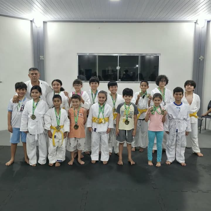 Fotografia de um grupo de judô onde um instrutor e várias crianças posam para uma foto após uma competição, tendo ao fundo janelas e uma moldura de espelho em uma parede branca e, em primeiro plano, o tatame preto e as crianças com medalhas e kimonos brancos. Fotografia de um grupo de judô onde um instrutor e várias crianças posam para uma foto após uma competição, tendo ao fundo janelas e uma moldura de espelho em uma parede branca e, em primeiro plano, o tatame preto e as crianças com medalhas e kimonos brancos.