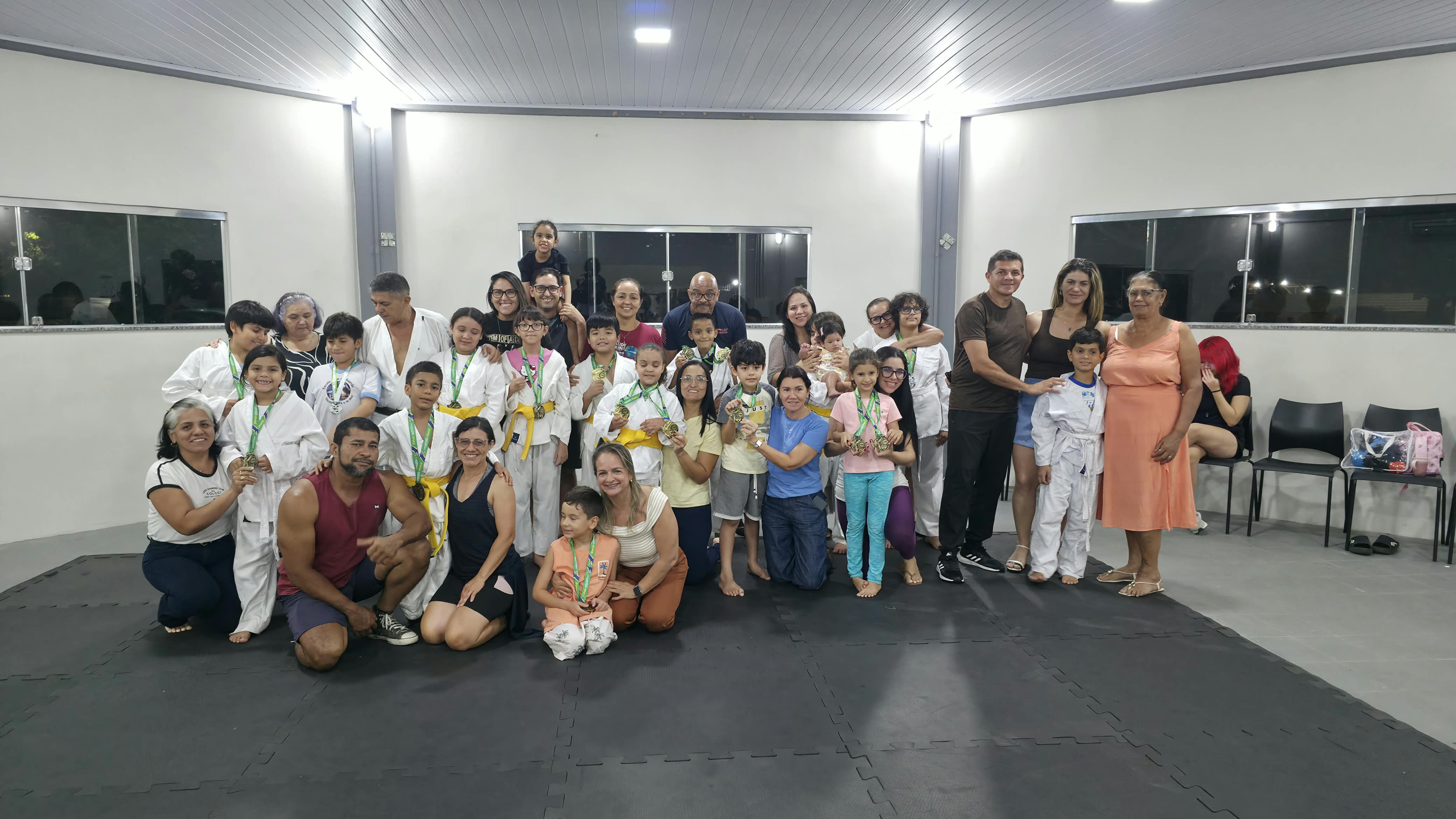 Fotografia de um grande grupo de famílias onde adultos e crianças, alguns em kimonos e outros com roupas casuais, posam juntos após um evento na academia de judô, tendo ao fundo janelas em uma parede branca e, em primeiro plano, o tatame preto e o grupo de pessoas ajoelhadas e em pé Fotografia de um grande grupo de famílias onde adultos e crianças, alguns em kimonos e outros com roupas casuais, posam juntos após um evento na academia de judô, tendo ao fundo janelas em uma parede branca e, em primeiro plano, o tatame preto e o grupo de pessoas ajoelhadas e em pé