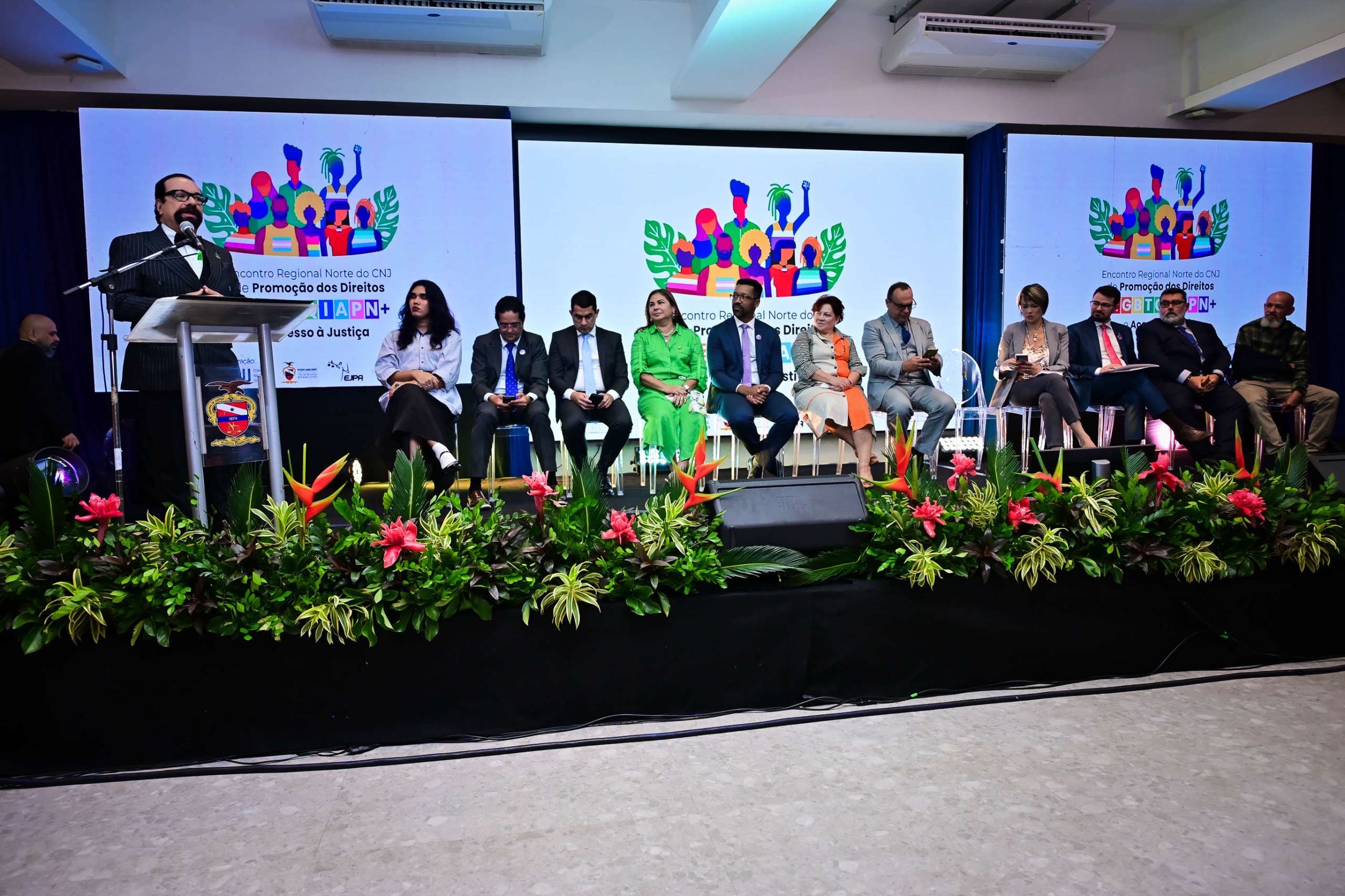 Imagem colorida mostra o palco do evento decorado com flores e um grande painel ao fundo. O presidente do TJRR, desembargador Leonardo Cupello, está em pé à esquerda, discursando em um púlpito com microfone. Ao seu lado, várias autoridades e convidados estão sentados em fileira acompanhando a apresentação. Imagem colorida mostra o palco do evento decorado com flores e um grande painel ao fundo. O presidente do TJRR, desembargador Leonardo Cupello, está em pé à esquerda, discursando em um púlpito com microfone. Ao seu lado, várias autoridades e convidados estão sentados em fileira acompanhando a apresentação.