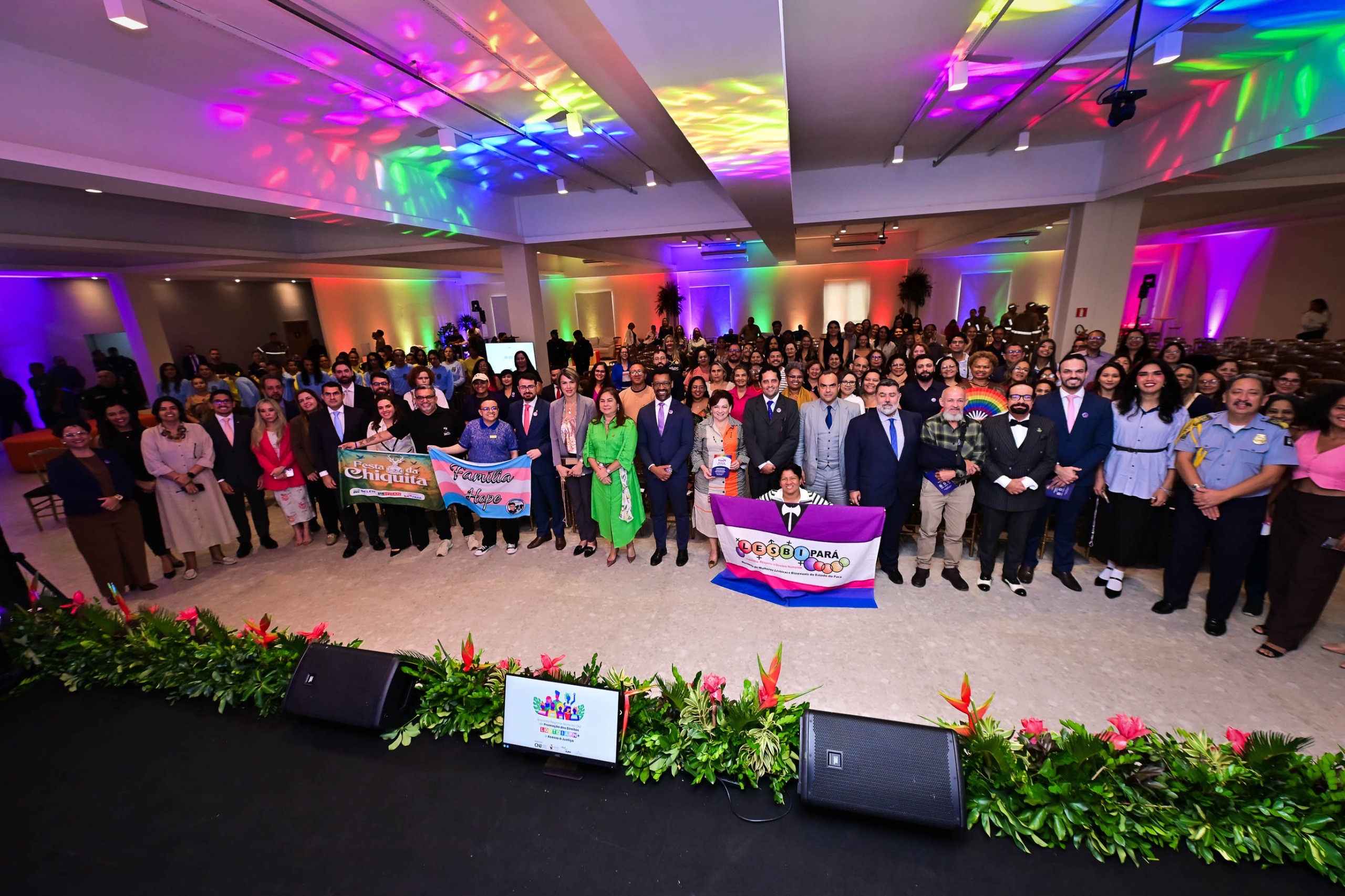 Imagem colorida mostra um grande grupo de pessoas reunidas em um salão amplo e iluminado com luzes coloridas, posando para uma foto oficial. Algumas pessoas na frente seguram faixas com mensagens de diversidade e representatividade. O presidente do TJRR, desembargador Leonardo Cupello, está posicionado ao centro-direita, vestindo terno risca de giz. Imagem colorida mostra um grande grupo de pessoas reunidas em um salão amplo e iluminado com luzes coloridas, posando para uma foto oficial. Algumas pessoas na frente seguram faixas com mensagens de diversidade e representatividade. O presidente do TJRR, desembargador Leonardo Cupello, está posicionado ao centro-direita, vestindo terno risca de giz.