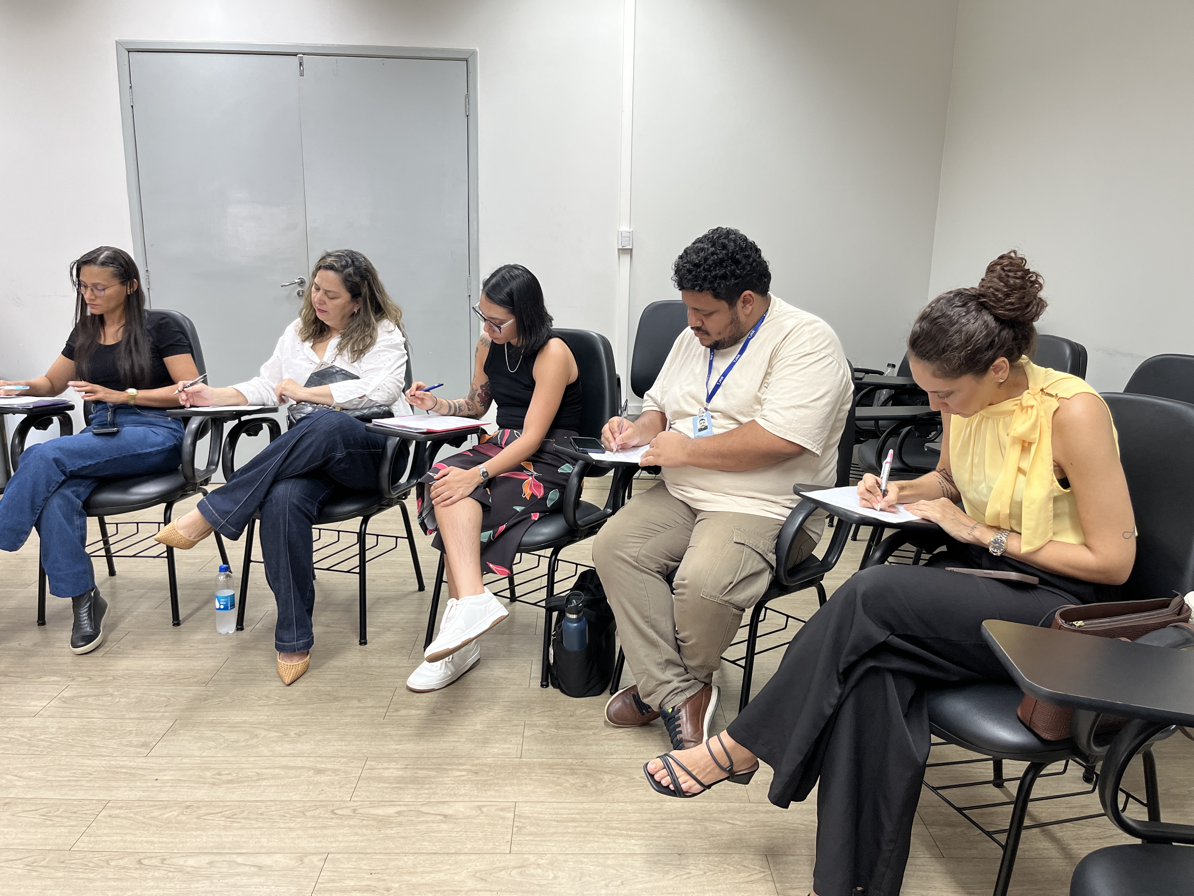 Imagem colorida mostra cinco participantes sentados lado a lado em uma sala de aula, preenchendo materiais impressos durante a atividade de encerramento da formação. Eles aparecem concentrados, escrevendo nas folhas apoiadas nas pranchetas das cadeiras. Imagem colorida mostra cinco participantes sentados lado a lado em uma sala de aula, preenchendo materiais impressos durante a atividade de encerramento da formação. Eles aparecem concentrados, escrevendo nas folhas apoiadas nas pranchetas das cadeiras.