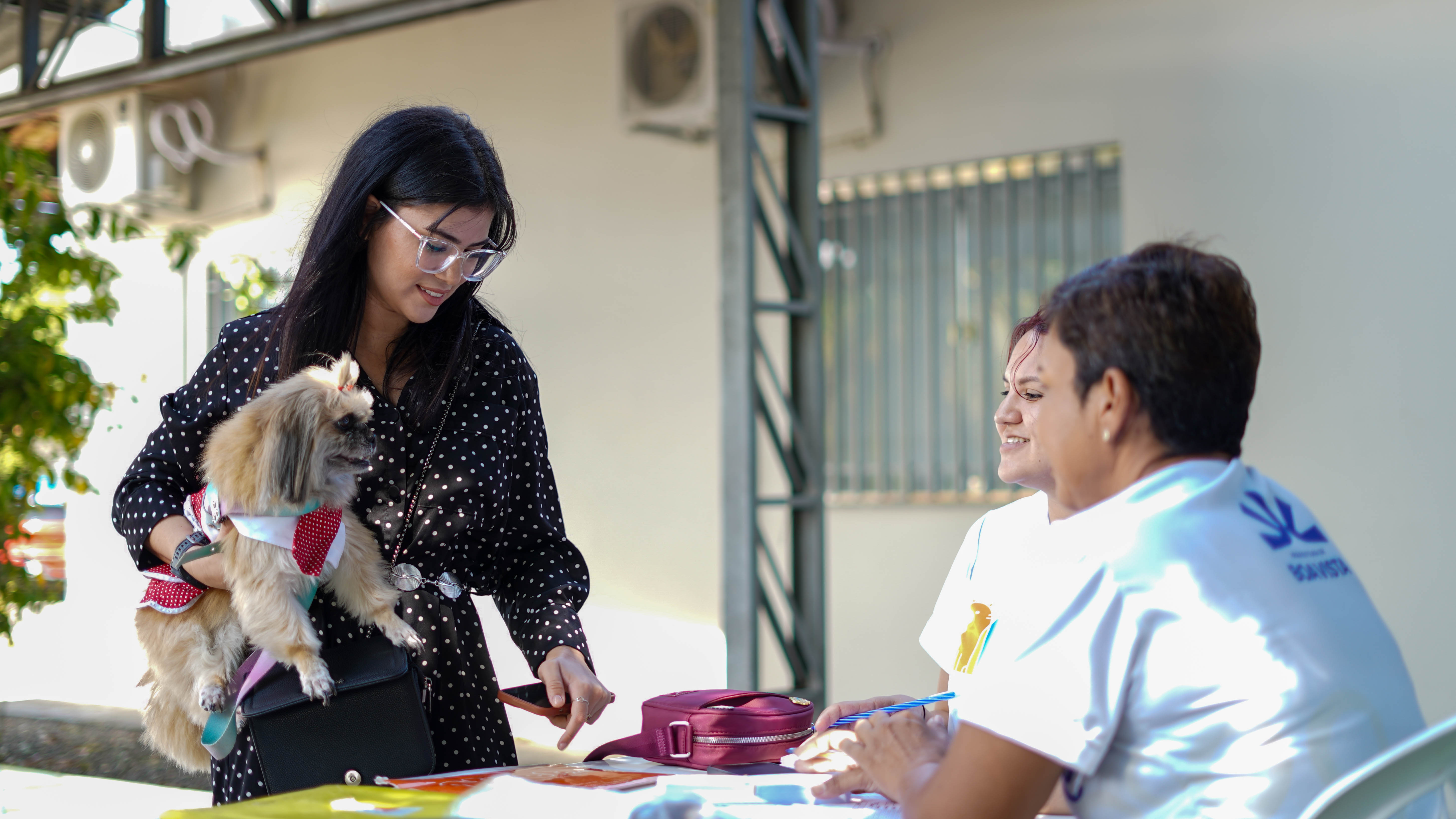 Mulher segura um cachorro pequeno enquanto conversa com uma profissional que está sentada à mesa, durante uma ação de atendimento. Mulher segura um cachorro pequeno enquanto conversa com uma profissional que está sentada à mesa, durante uma ação de atendimento.