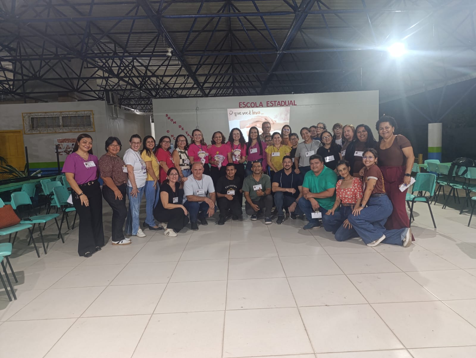 Fotografia em plano geral do grupo de participantes e organizadores do programa Justiça Comunitária reunidos em pé, sorrindo para o registro fotográfico no centro do salão coberto, com uma projeção institucional ao fundo.