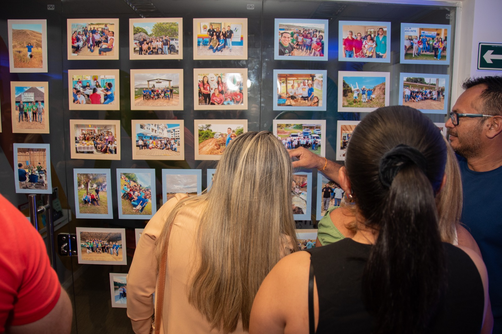Imagem colorida mostra pessoas observando um painel com fotografias de equipes, atendimentos e visitas a comunidades. Os registros estão organizados em fileiras, com molduras claras e azuis. À direita, um homem de óculos aponta para uma das fotos. Imagem colorida mostra pessoas observando um painel com fotografias de equipes, atendimentos e visitas a comunidades. Os registros estão organizados em fileiras, com molduras claras e azuis. À direita, um homem de óculos aponta para uma das fotos.