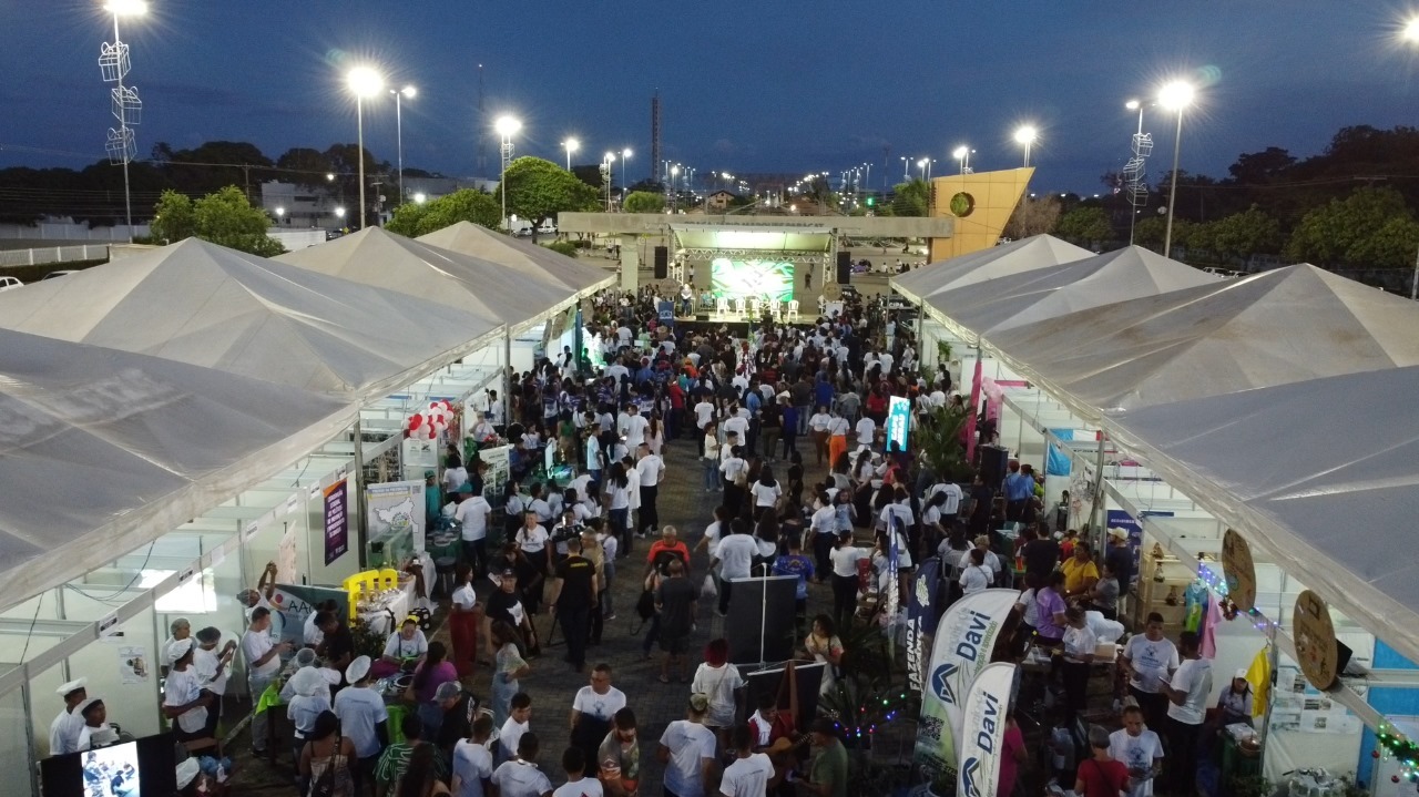 Imagem colorida, em vista aérea, mostra uma edição da Feira de Resultados dos Projetos Sociais no estacionamento da Praça Fábio Marques Paracat, durante o início da noite. Duas fileiras de tendas brancas abrigam estandes dos projetos, enquanto um grande público circula pelo espaço. Ao fundo, um palco iluminado concentra parte da programação do evento. Imagem colorida, em vista aérea, mostra uma edição da Feira de Resultados dos Projetos Sociais no estacionamento da Praça Fábio Marques Paracat, durante o início da noite. Duas fileiras de tendas brancas abrigam estandes dos projetos, enquanto um grande público circula pelo espaço. Ao fundo, um palco iluminado concentra parte da programação do evento.