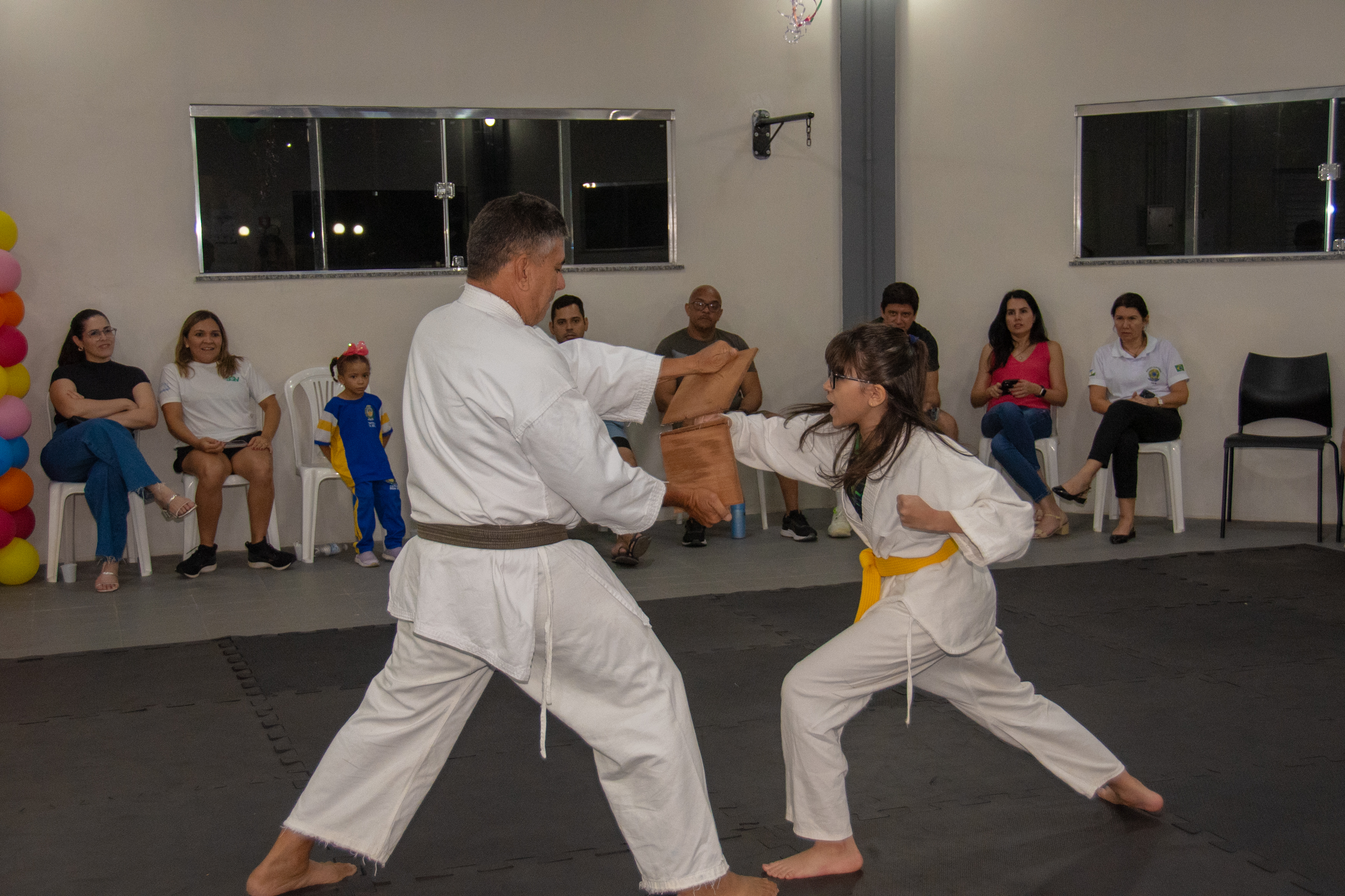 Imagem colorida mostra uma criança de quimono branco e faixa amarela treinando artes marciais com um instrutor em um tatame. Várias pessoas assistem à aula sentadas em cadeiras ao fundo, em uma sala iluminada. Imagem colorida mostra uma criança de quimono branco e faixa amarela treinando artes marciais com um instrutor em um tatame. Várias pessoas assistem à aula sentadas em cadeiras ao fundo, em uma sala iluminada.